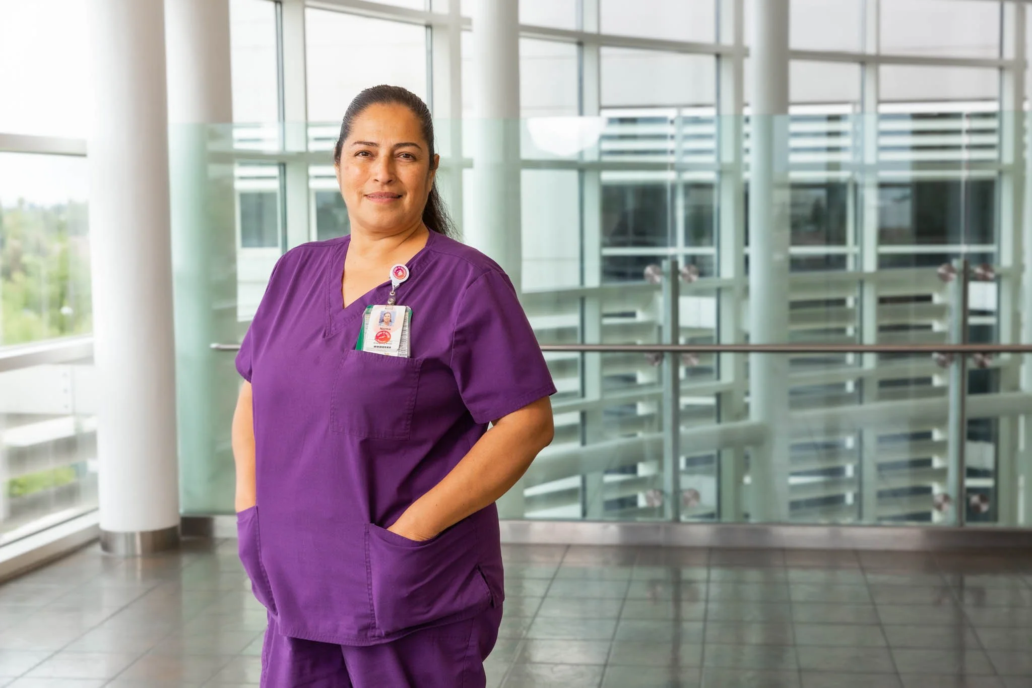 A female healthcare professional in purple scrubs standing in a modern hospital lobby with glass railings and large windows, smiling at the camera.