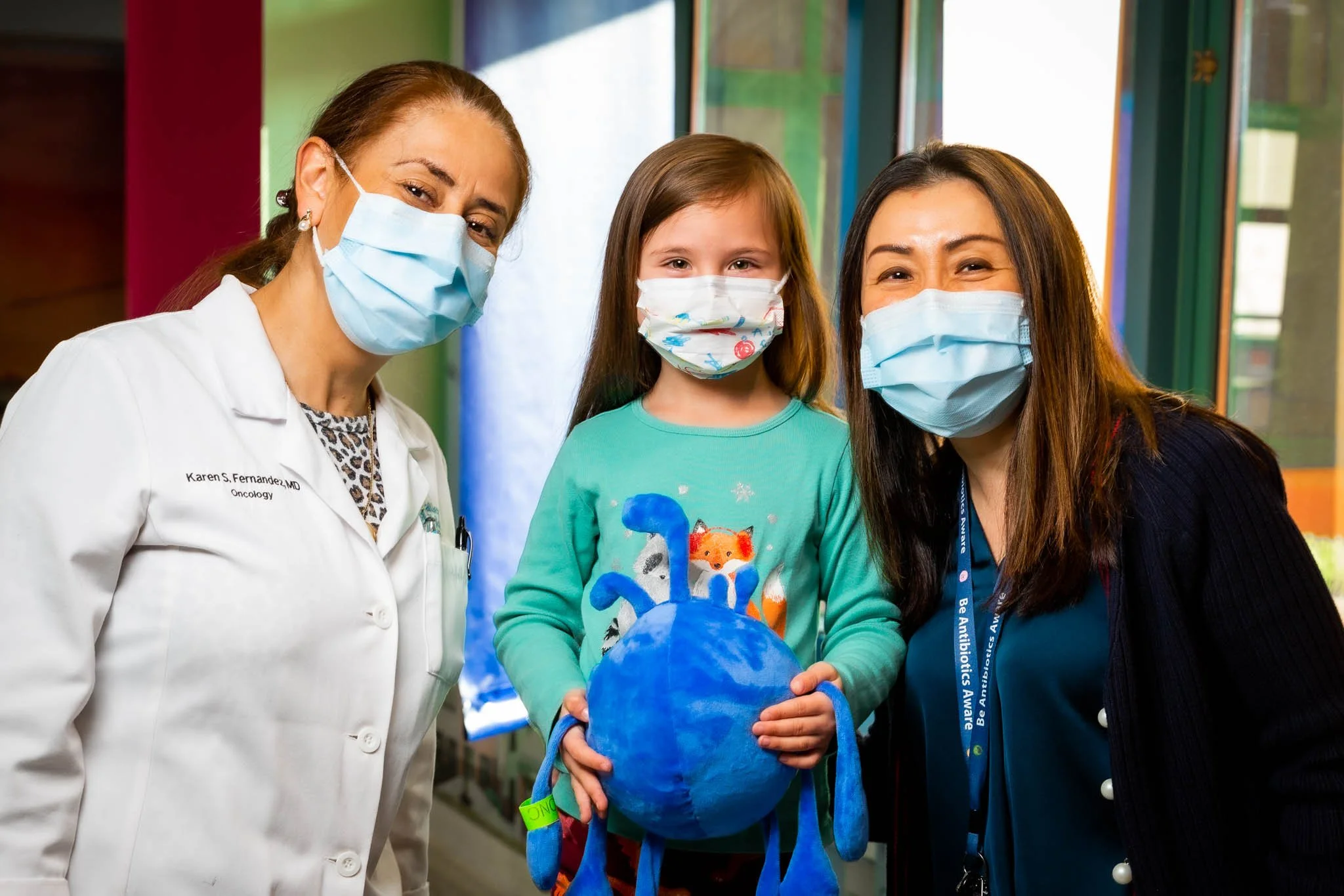 Three women and a young girl wearing masks, standing together indoors, with a colorful background. The young girl holds a blue craft globe.