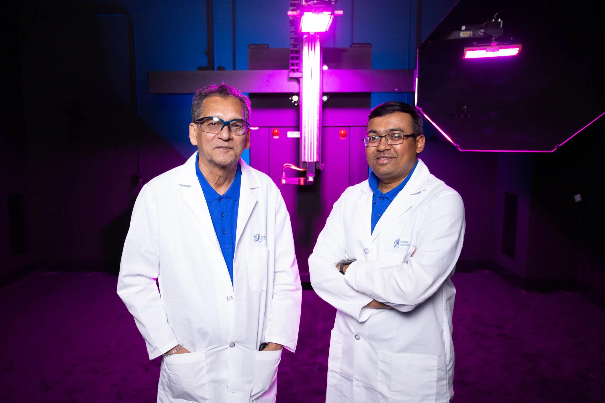 Two scientists wearing white lab coats stand in a room with advanced technological equipment and purple and pink lighting.