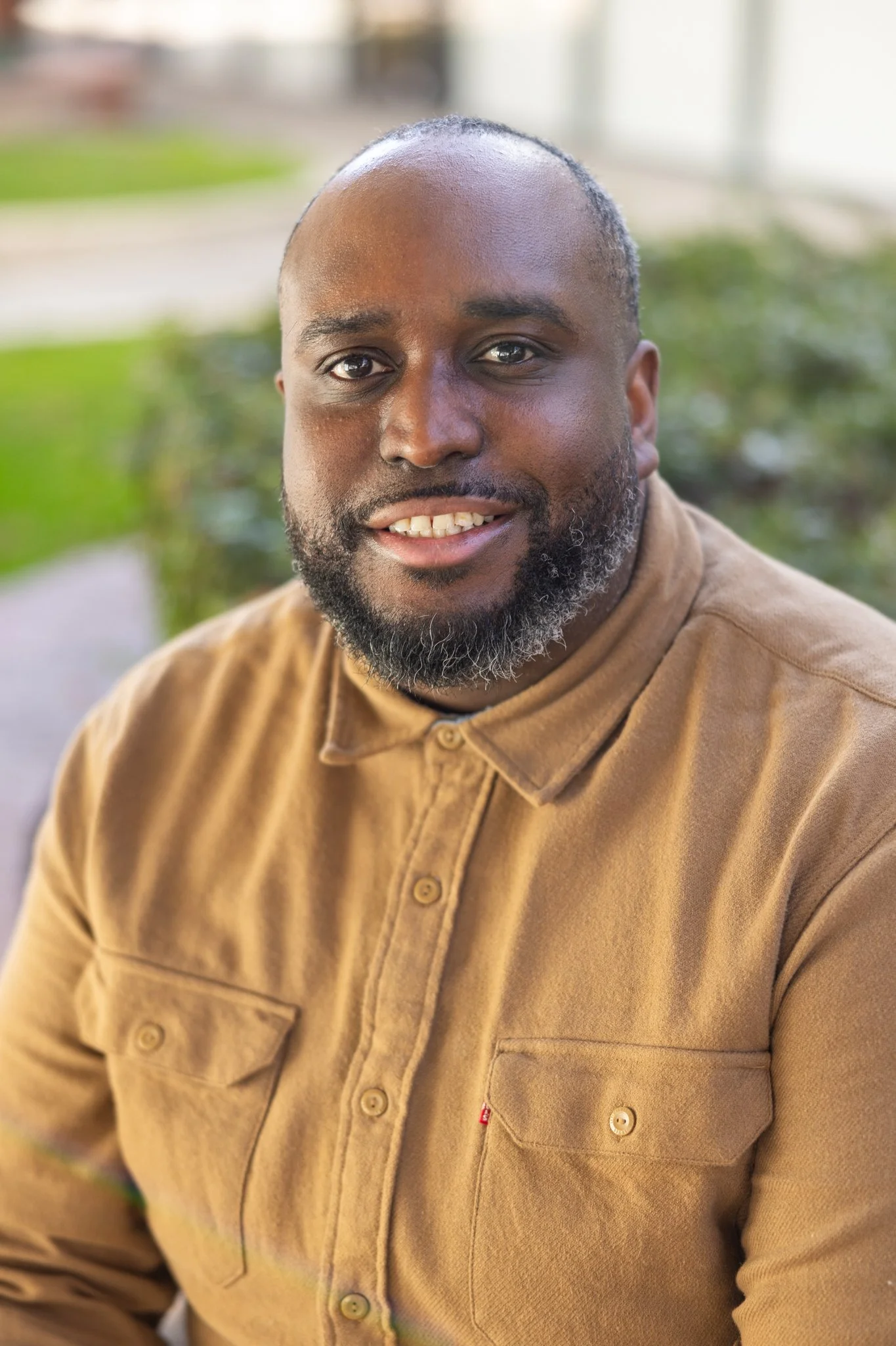 Close-up portrait of a smiling Black man with a beard, wearing a tan button-up shirt, sitting outdoors with blurred greenery in the background.