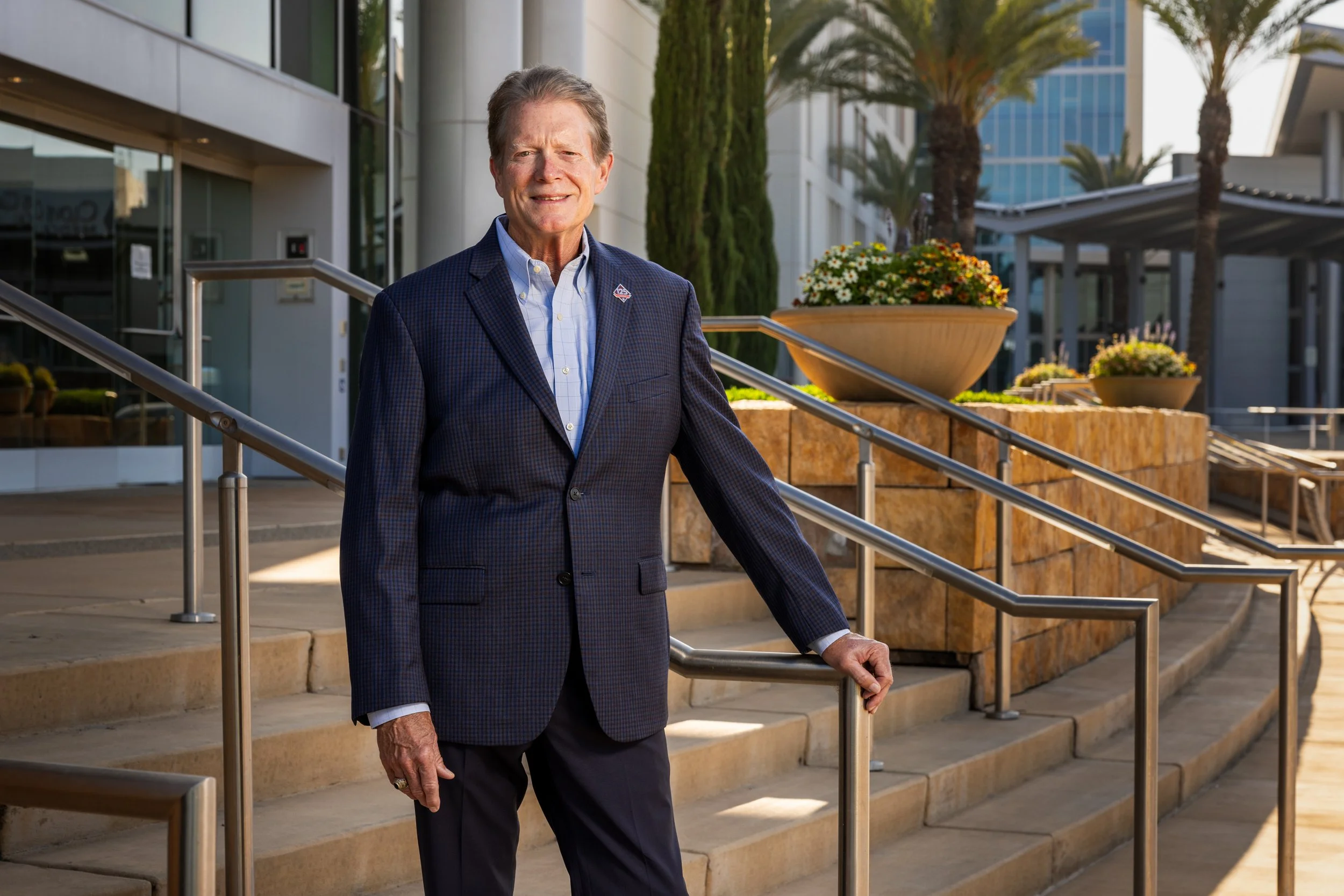 A middle-aged man in a business suit standing outside of a modern building holding a railing. The background features a landscaped area with large flower pots, lush greenery, and tall palm trees. The weather appears sunny and bright.