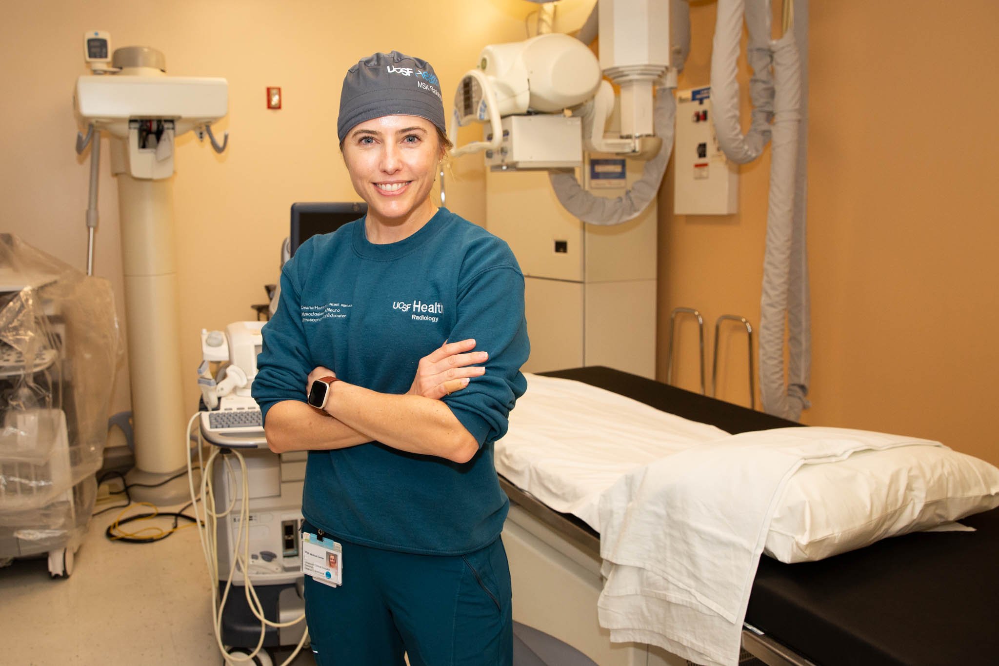 A smiling woman wearing scrubs and a surgical cap standing in a hospital room.