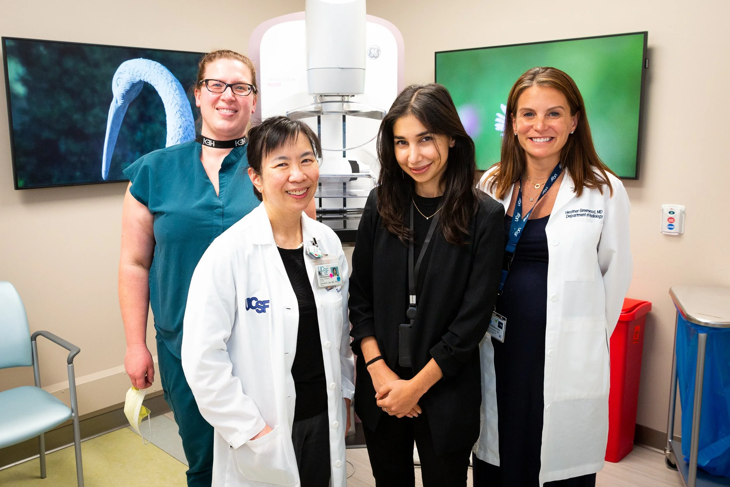 Group of four women in medical attire posing inside a hospital or clinic room with medical equipment and monitors in the background.
