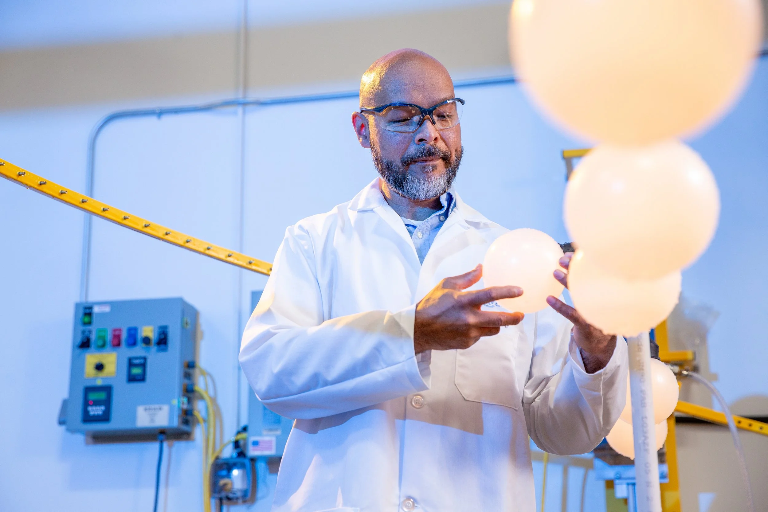 A scientist in a white lab coat and safety glasses examining illuminated globes in a laboratory setting.
