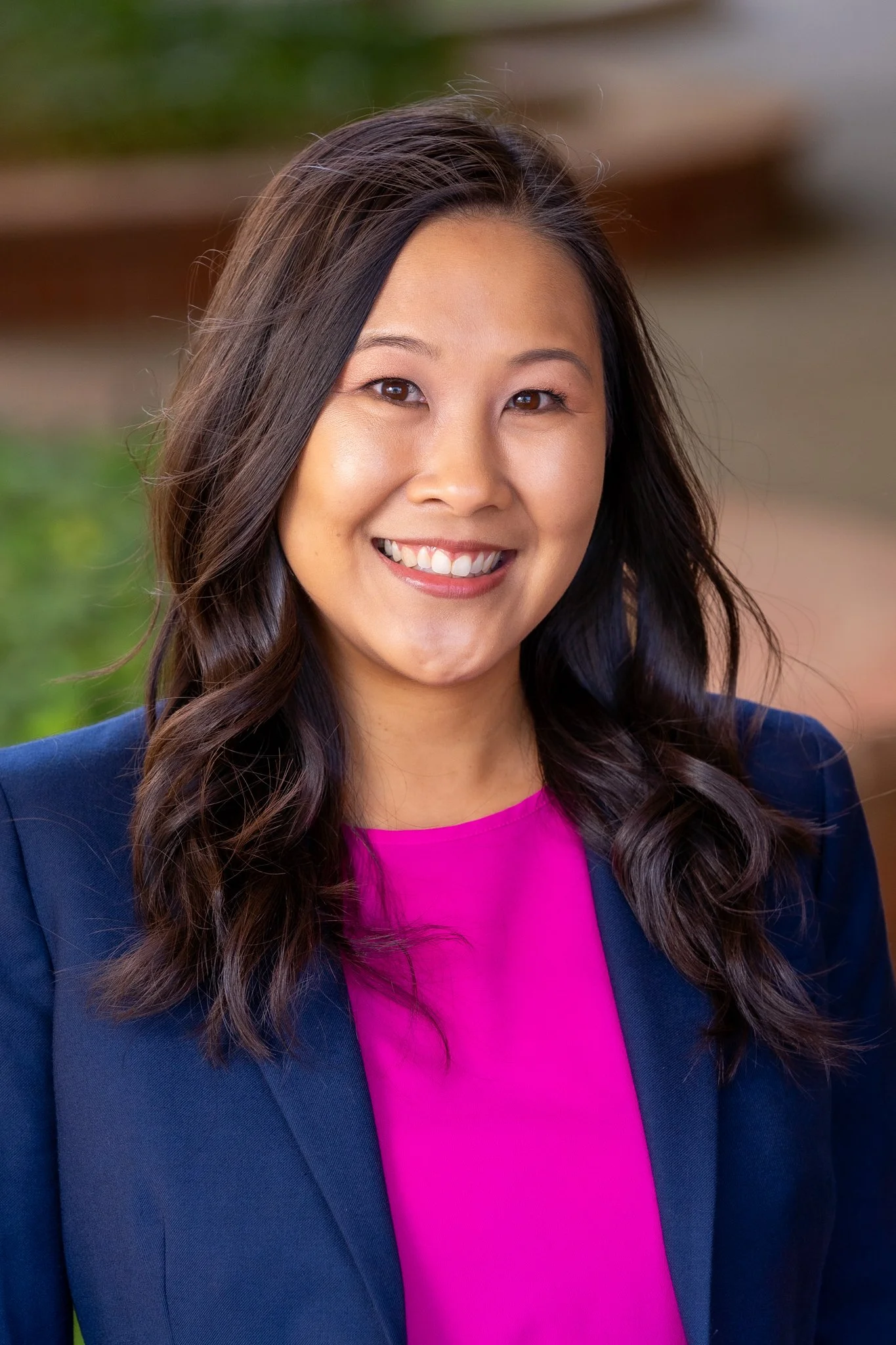 Smiling woman with long dark hair wearing a navy blazer and a bright pink blouse, standing outdoors.