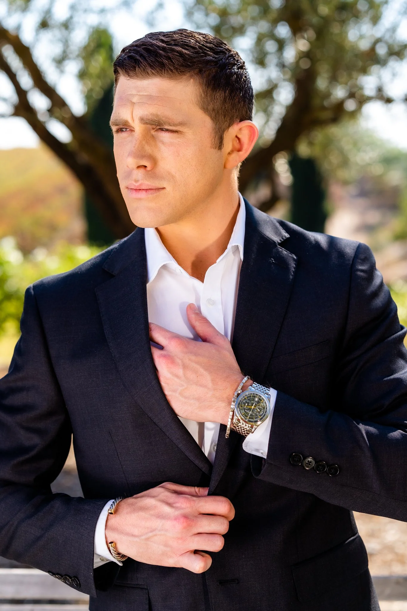 A man in a black suit with a white shirt, wearing a wristwatch and bracelet, is touching his chest with one hand and holding his jacket with the other. He is outdoors with blurred trees in the background.