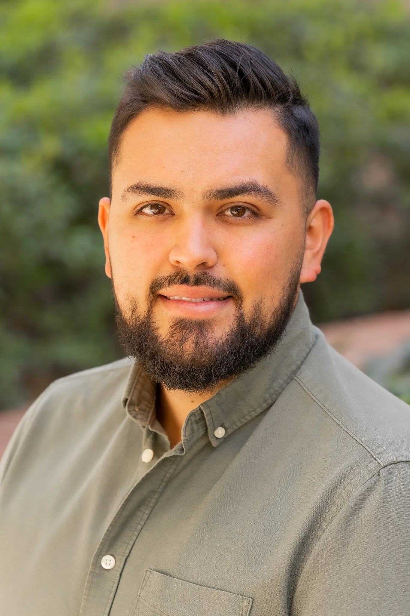 Close-up portrait of a young man with dark hair, beard, and mustache, wearing a gray button-up shirt, outdoors with blurred greenery in the background.