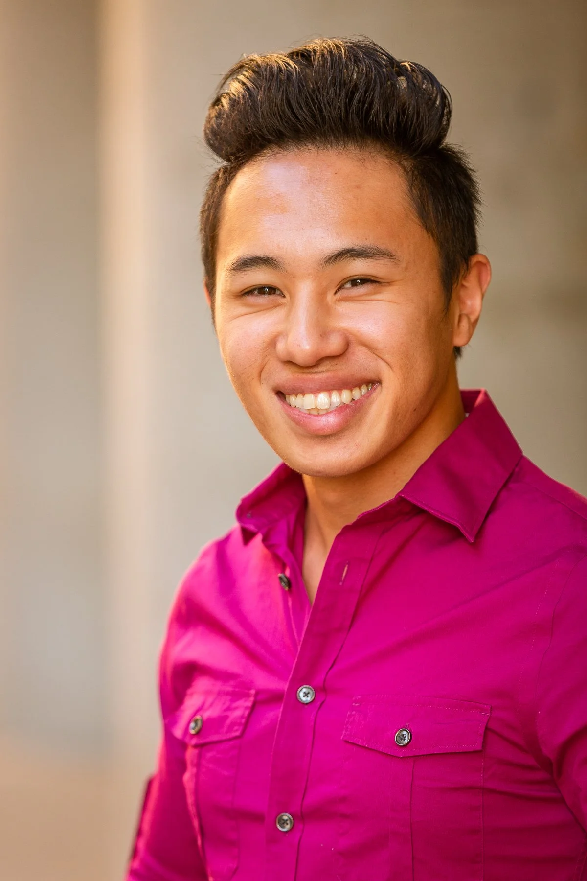 A young man smiling, wearing a bright magenta shirt, with short black hair styled upwards, standing in front of a blurred background.