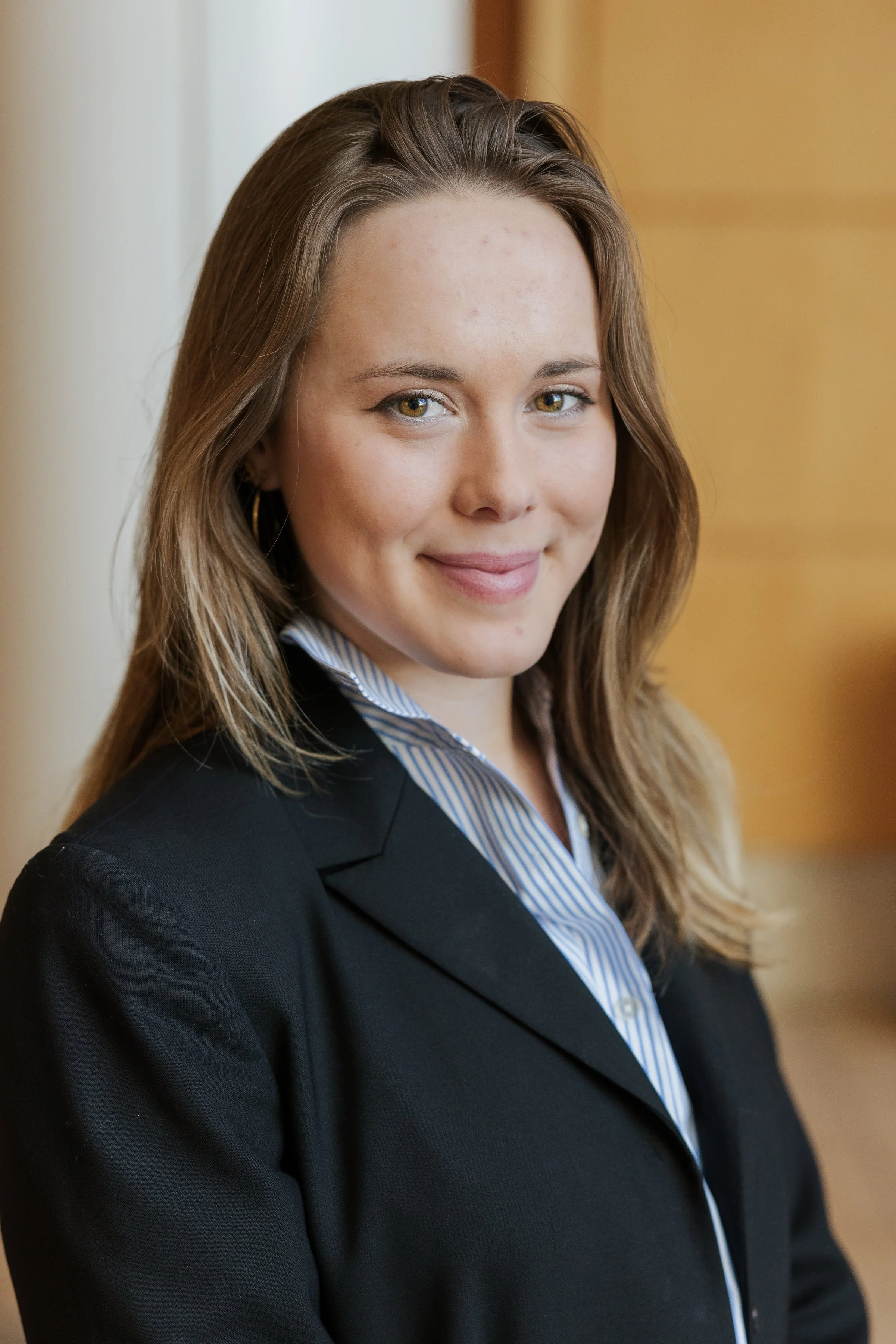 A young woman with shoulder-length light brown hair, wearing a black blazer and a blue and white striped collared shirt, smiling at the camera in an indoor setting.