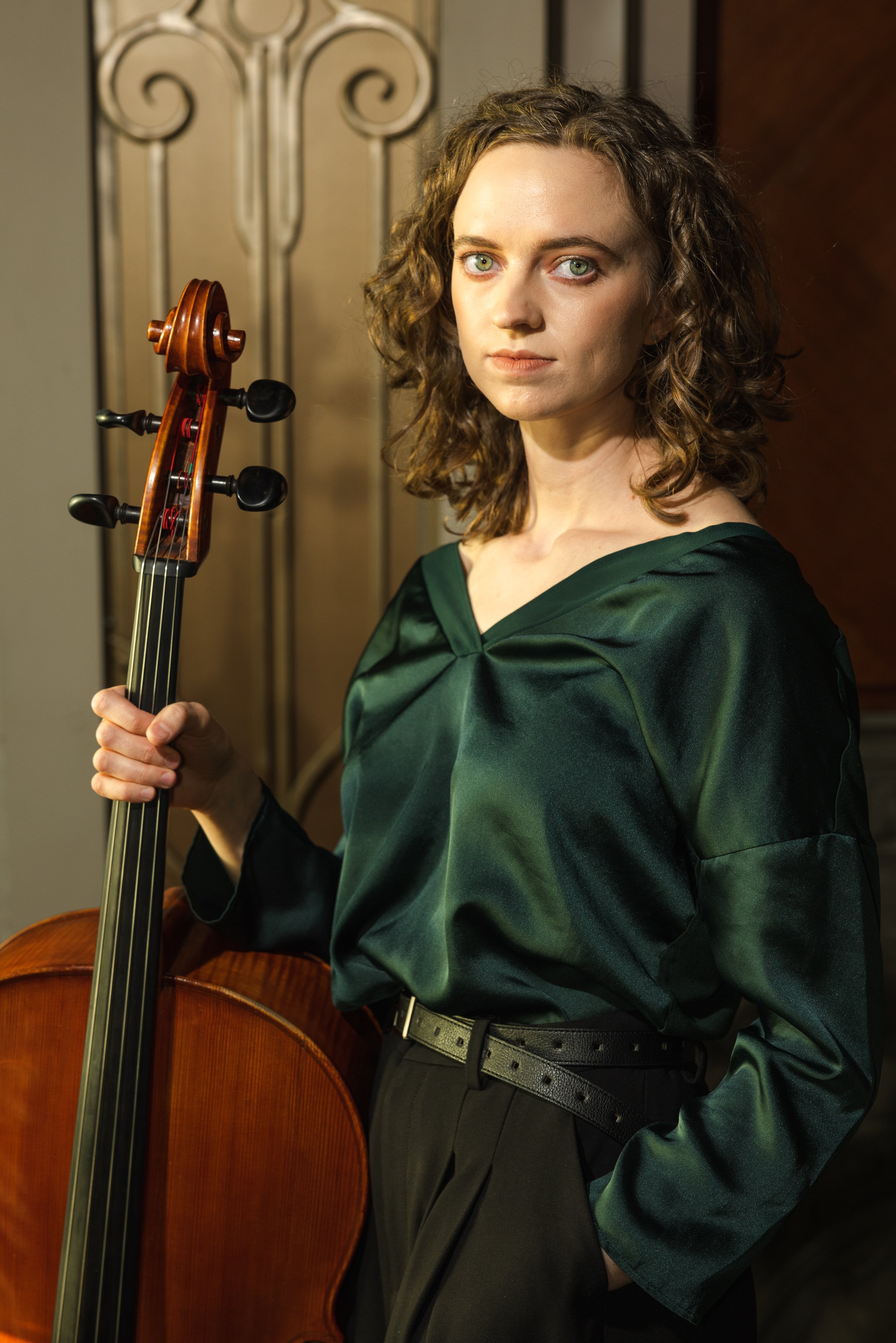 A woman with curly hair holding a cello, standing indoors near a decorative door, wearing a dark green satin blouse and black pants.
