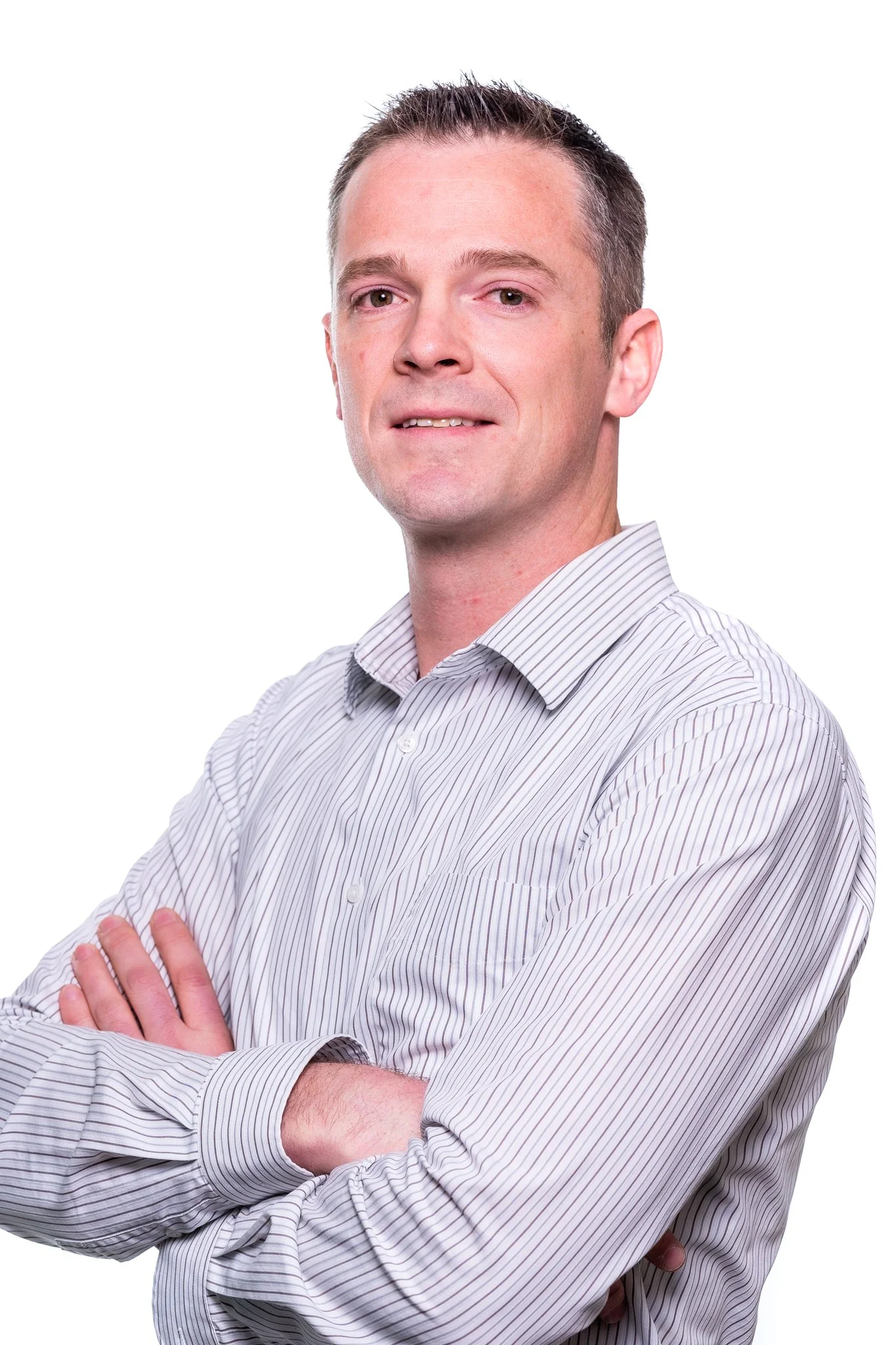 A man with short, dark hair and fair skin wearing a light-colored, striped button-up shirt, standing with arms crossed and a slight smile against a plain white background.
