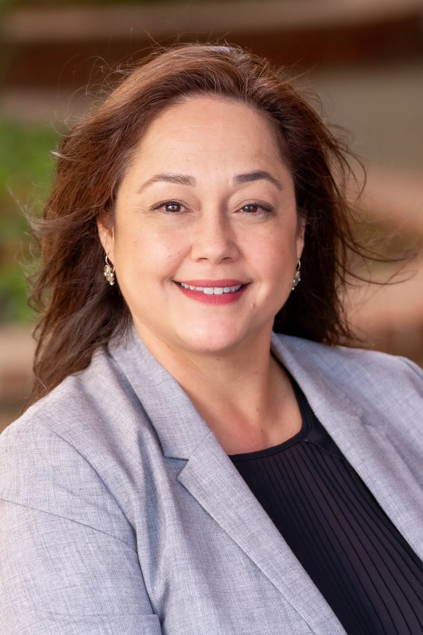Portrait of a smiling middle-aged woman with brown hair, small earrings, wearing a light gray blazer and black top, outdoor background.