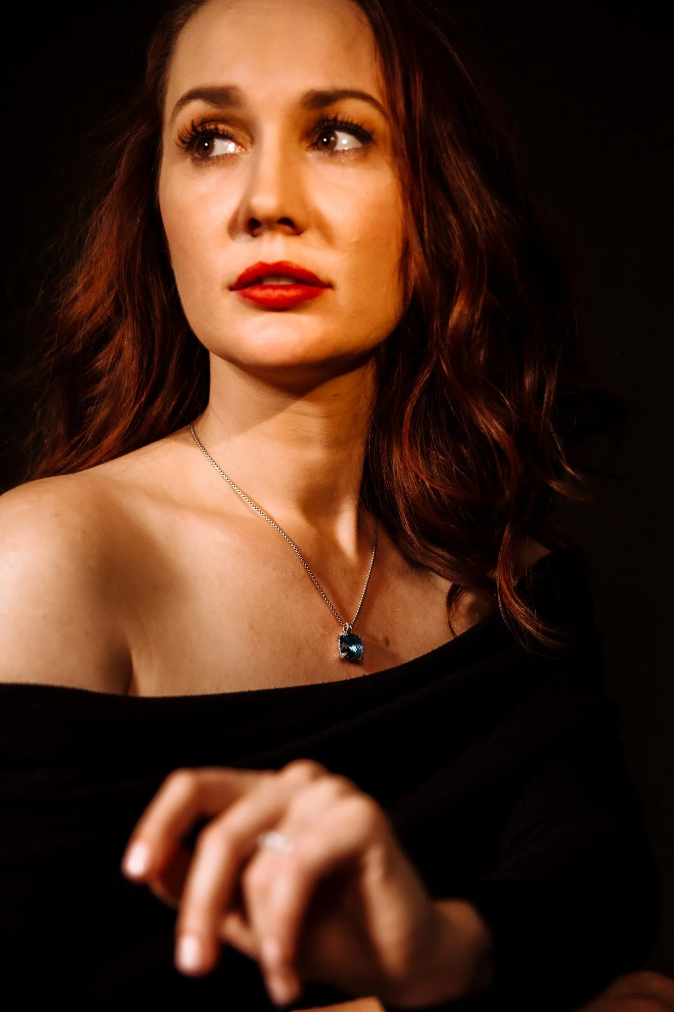 A woman with auburn hair and red lipstick wearing an off-the-shoulder black top and a necklace with a blue gemstone pendant.
