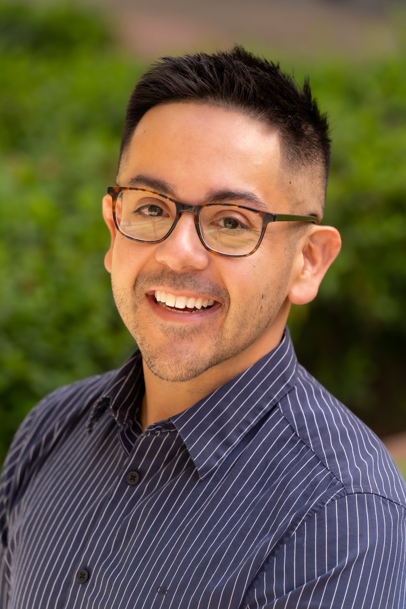 Close-up portrait of a smiling man with black hair, glasses, wearing a navy blue striped shirt, outdoors with blurred green foliage background.