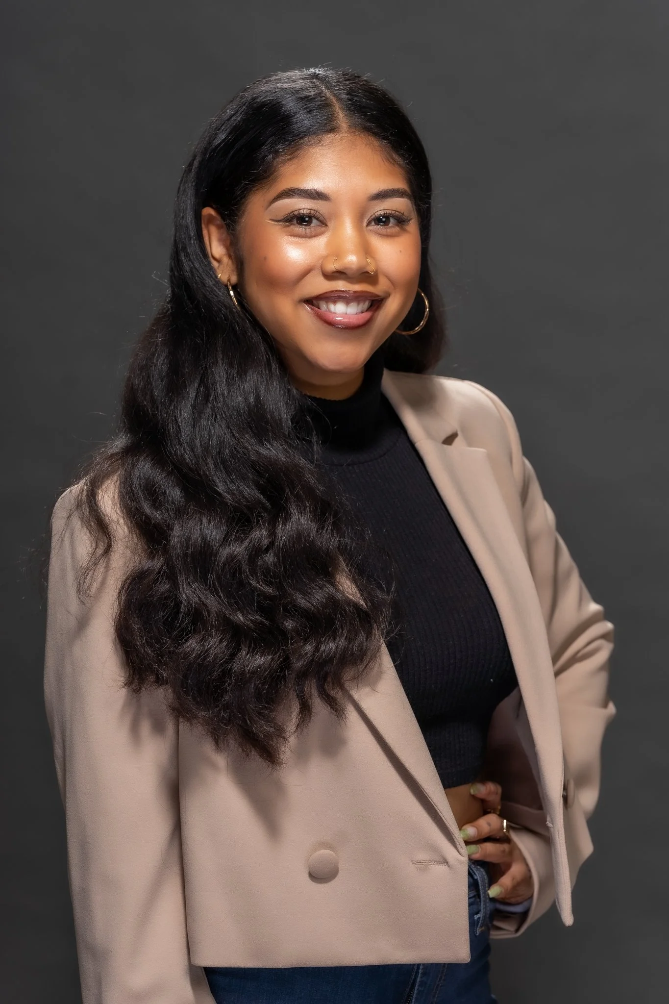 Portrait of a woman with long dark wavy hair, smiling, wearing a beige blazer over a black top, and gold hoop earrings, standing against a dark background.