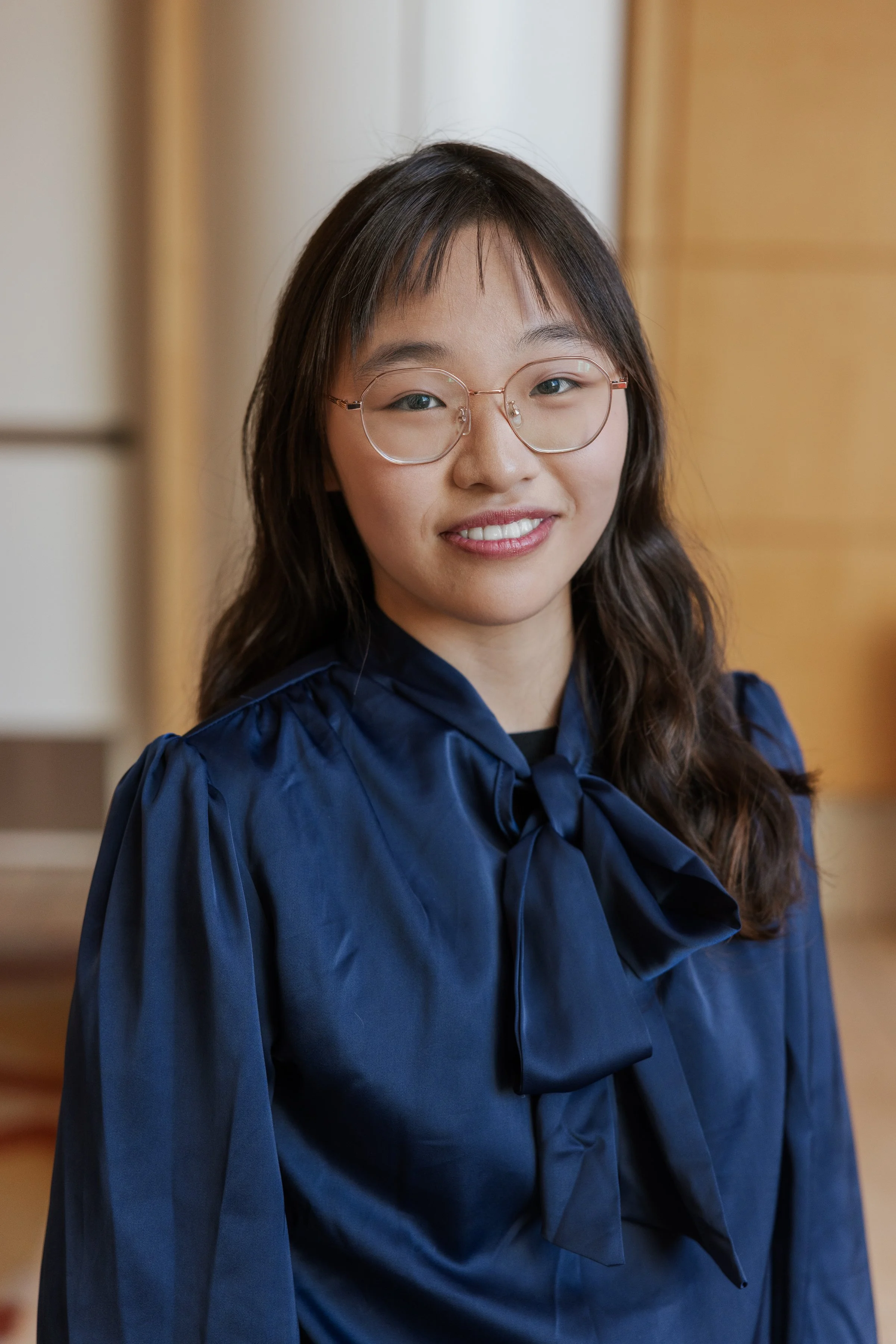 A young woman with glasses and wavy hair wearing a blue dress with a bow, smiling in an indoor setting.