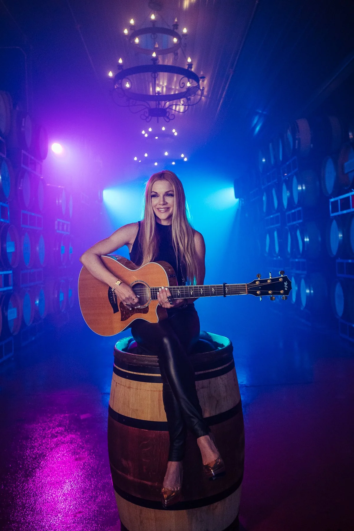 Woman with long blonde hair sitting on a wine barrel, playing an acoustic guitar, in a room with blue and purple lighting, wine barrel walls, and chandeliers hanging from the ceiling.