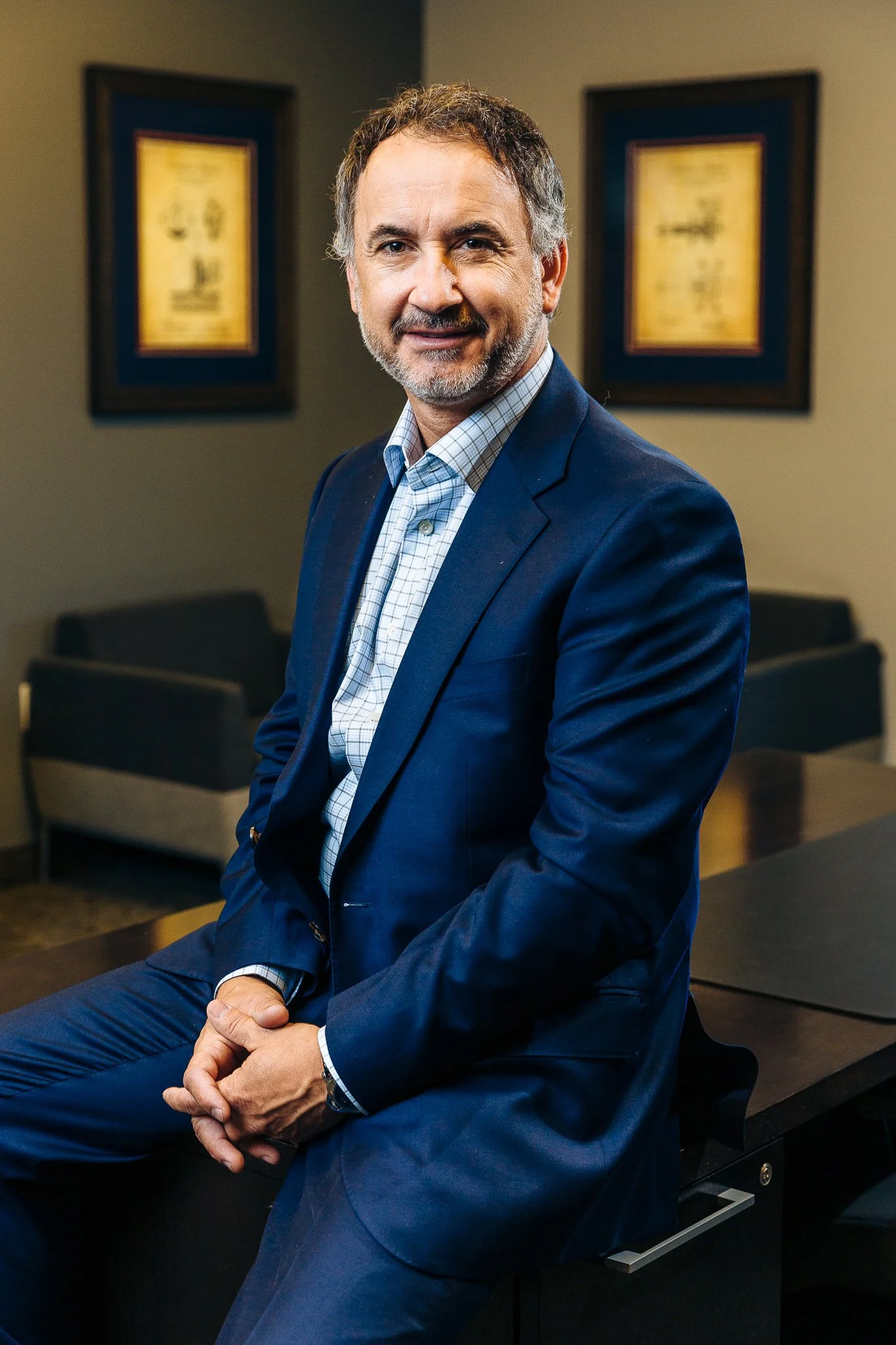 A middle-aged man with a beard, wearing a navy blue suit and checkered shirt, sitting on the edge of a conference table in a professional office setting.