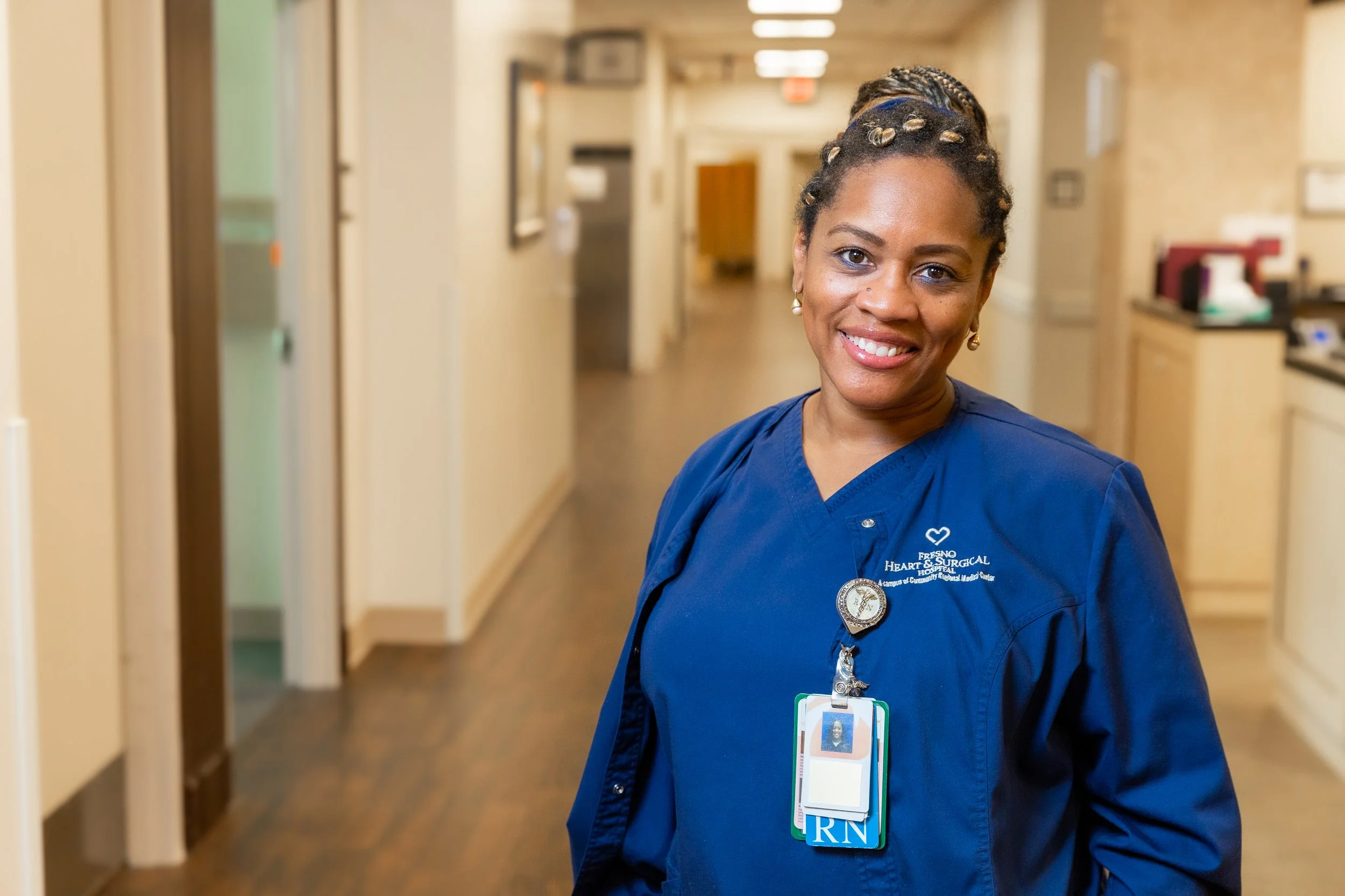 A smiling nurse wearing navy scrubs with a badge, standing in a hospital corridor.