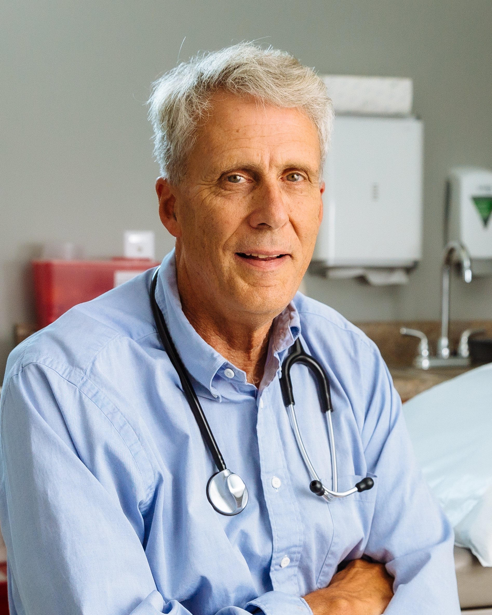 A male doctor with gray hair wearing a light blue shirt and stethoscope, sitting in a medical office.