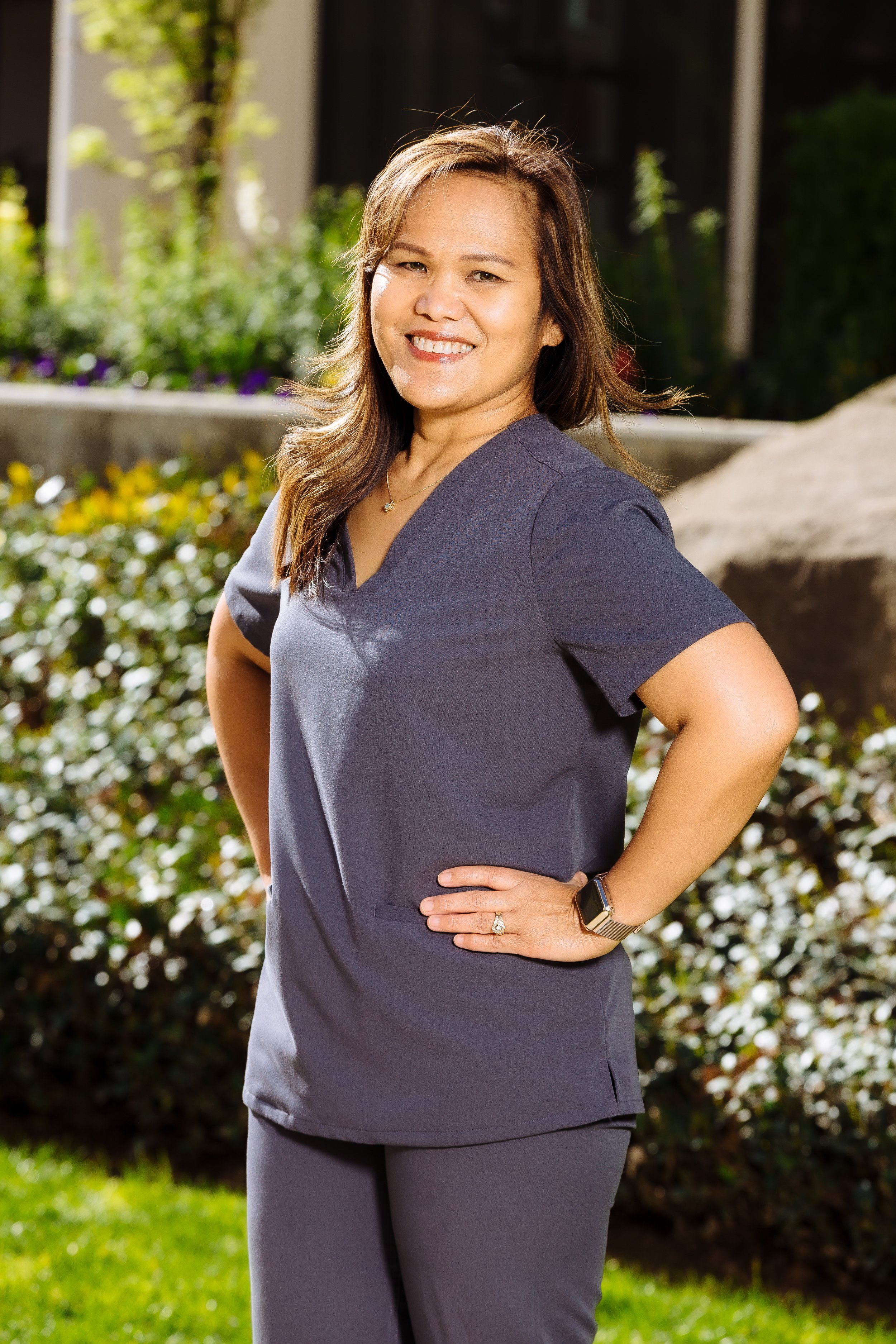 A woman wearing navy medical scrubs standing outdoors with her hand on her hip, smiling, with greenery and a large rock in the background.