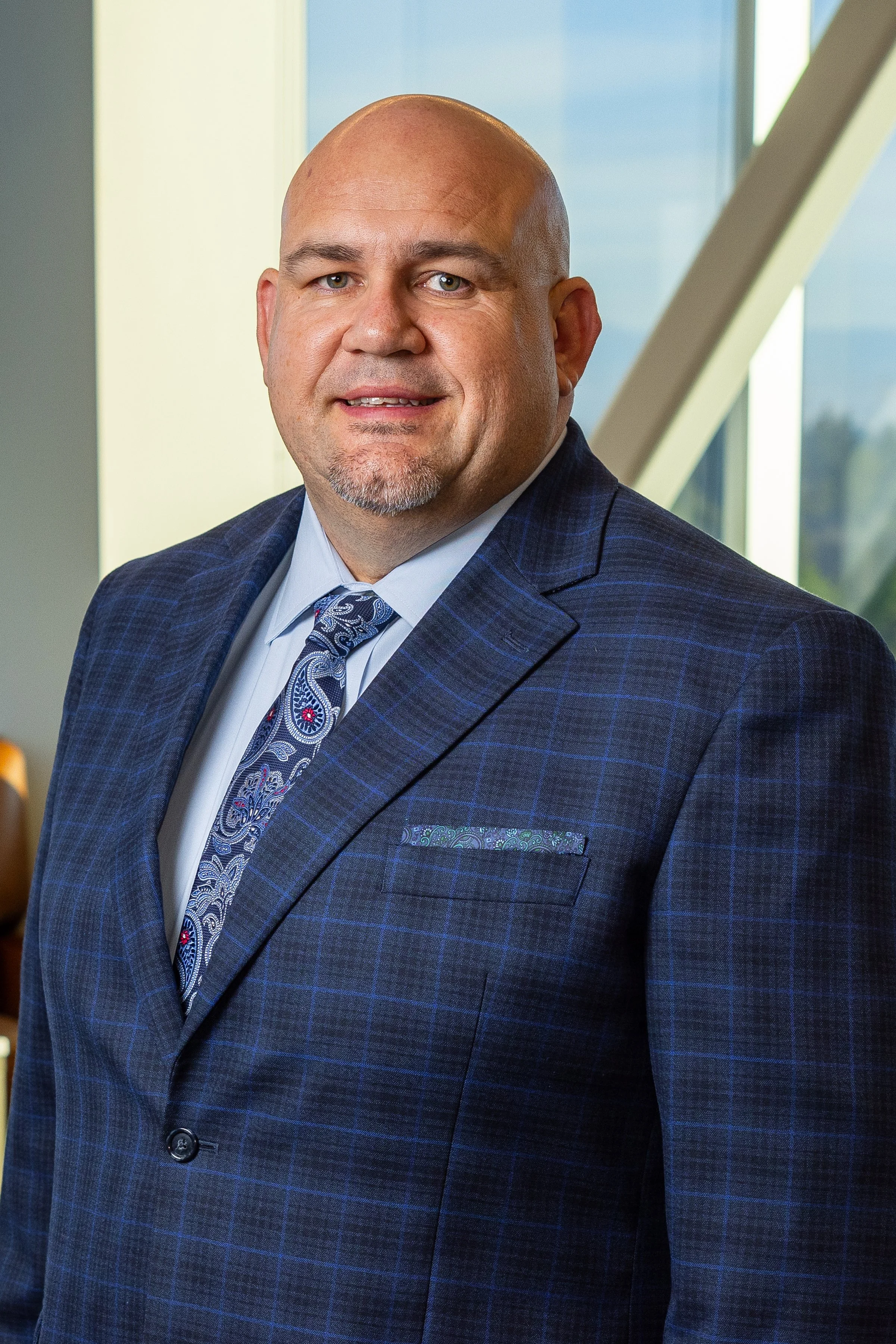 Professional man in a blue checkered suit and paisley tie, standing in an office with large windows.