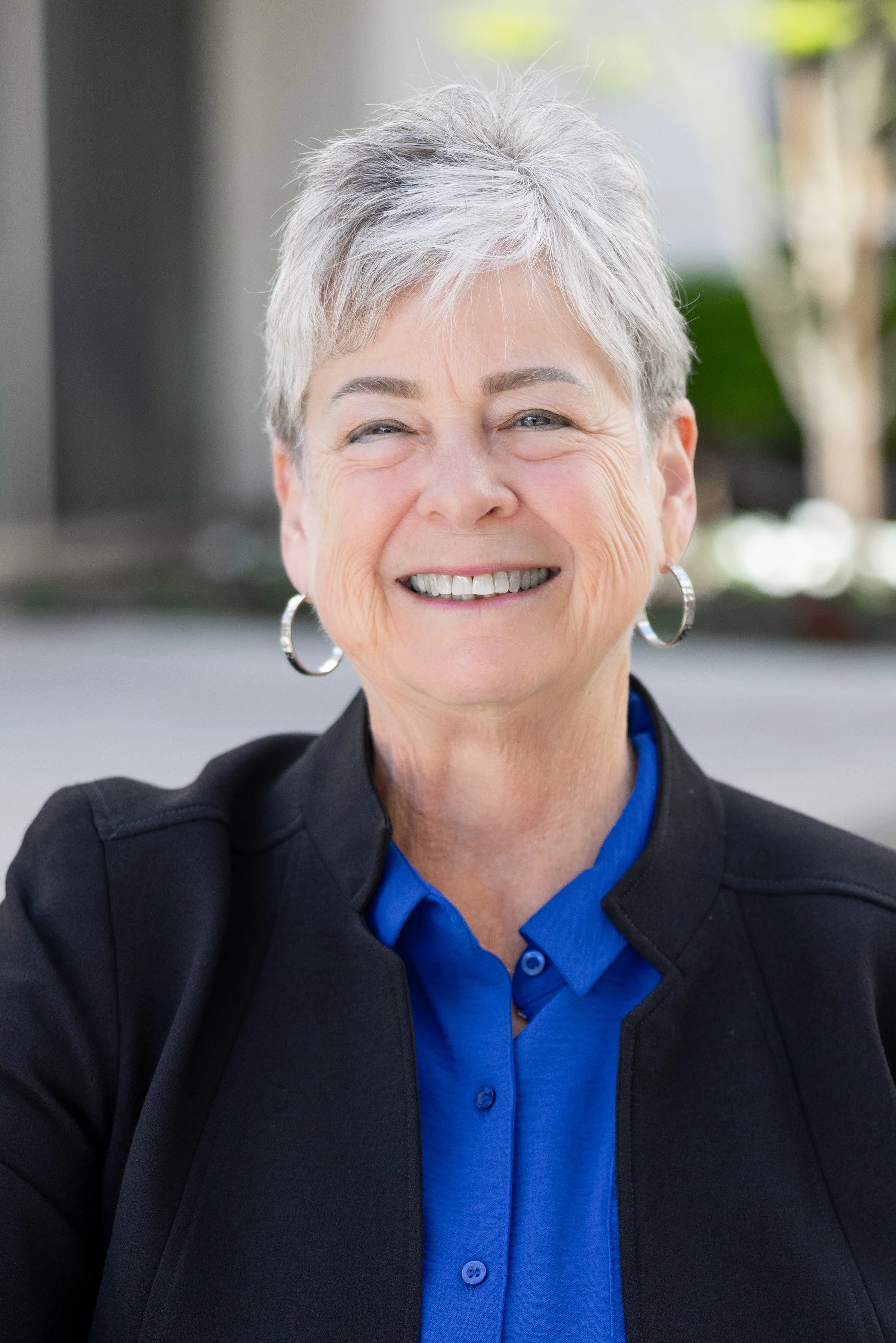 A smiling older woman with short gray hair, wearing a blue blouse, black jacket, and hoop earrings, outdoors.
