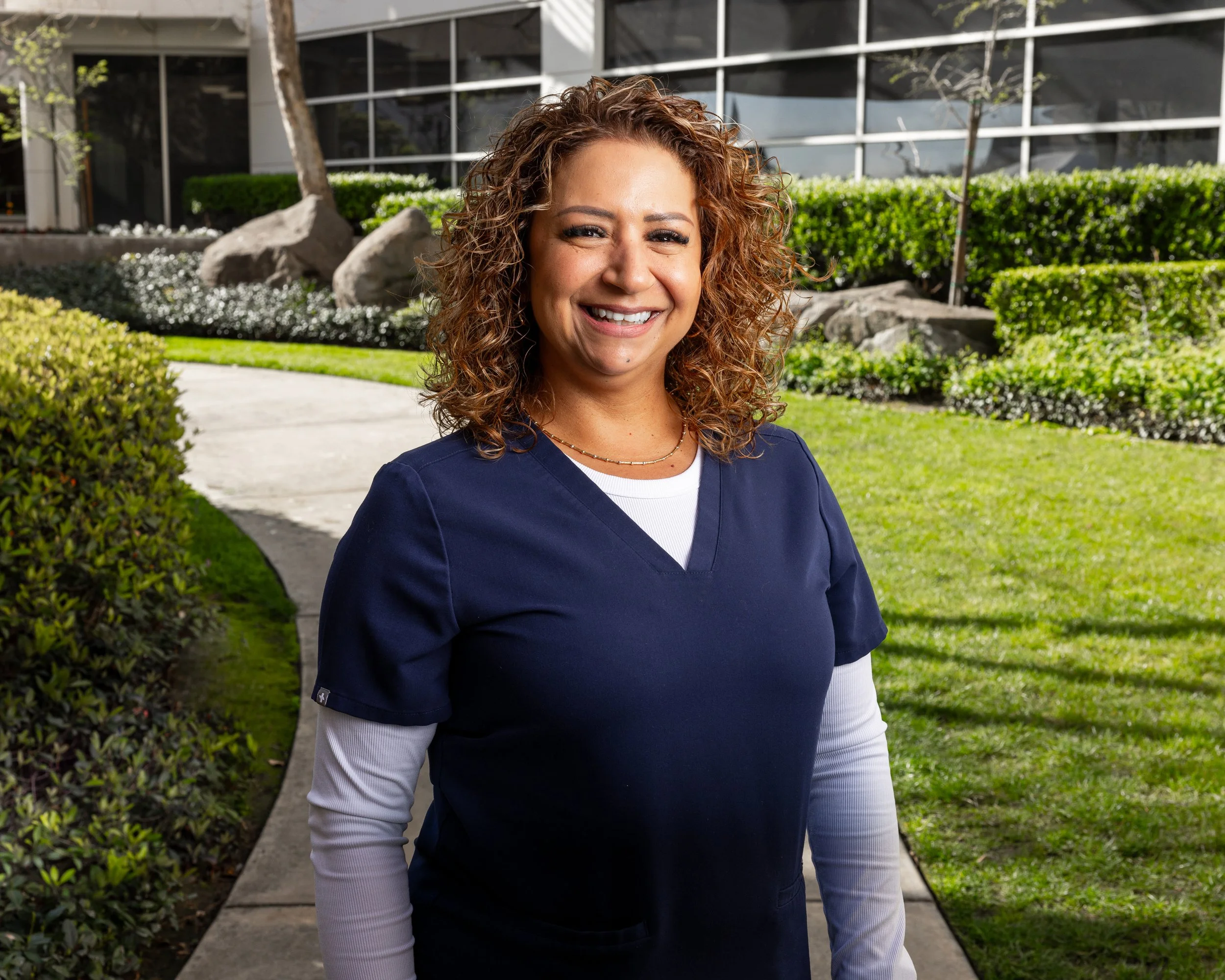A woman with curly hair wearing navy scrubs and a white long-sleeve shirt under her scrubs, smiling outdoors on a walkway with greenery and modern buildings in the background.