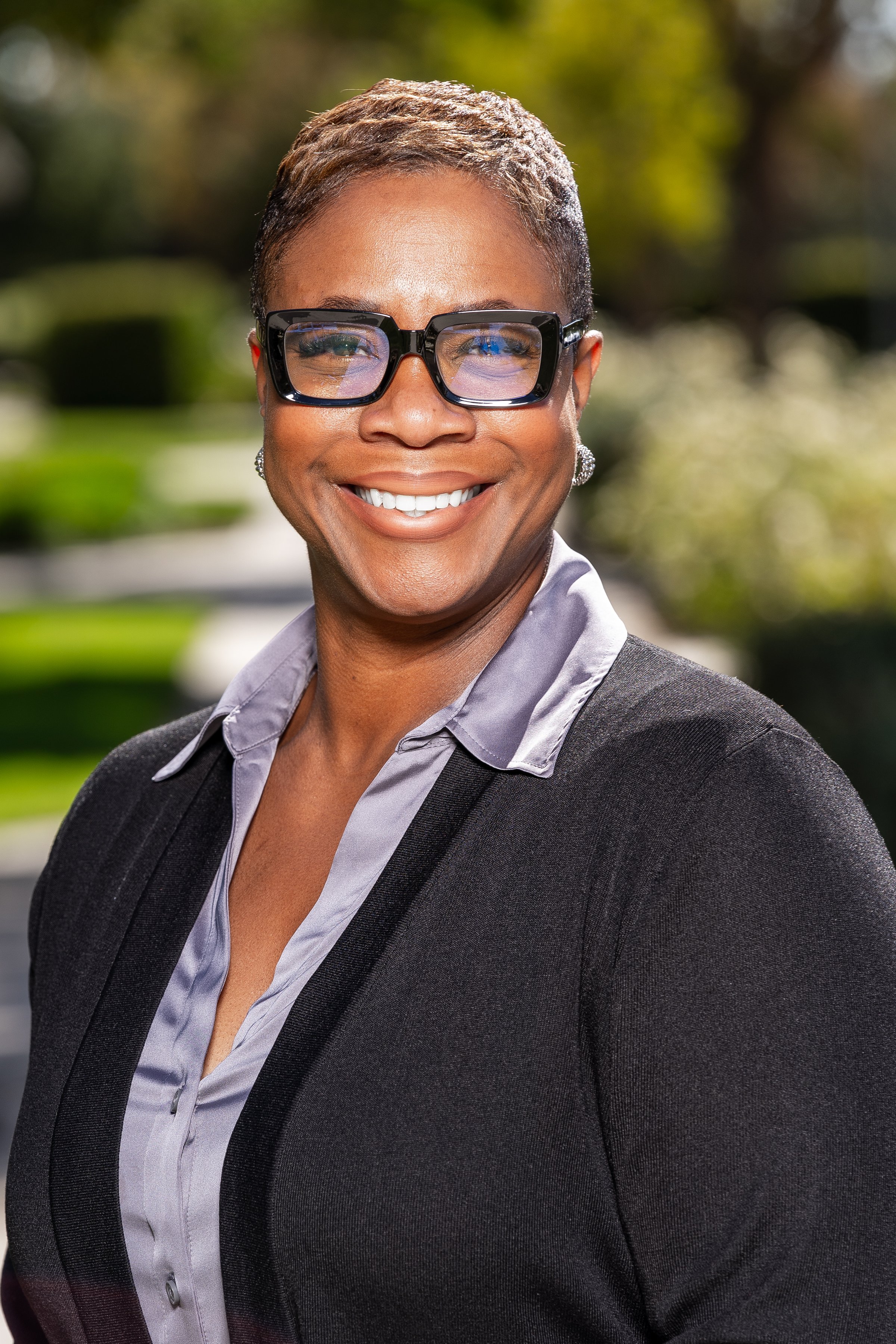 A professional woman with short, textured hair, wearing glasses, a gray blouse, and a black blazer, smiling outdoors with greenery in the background.