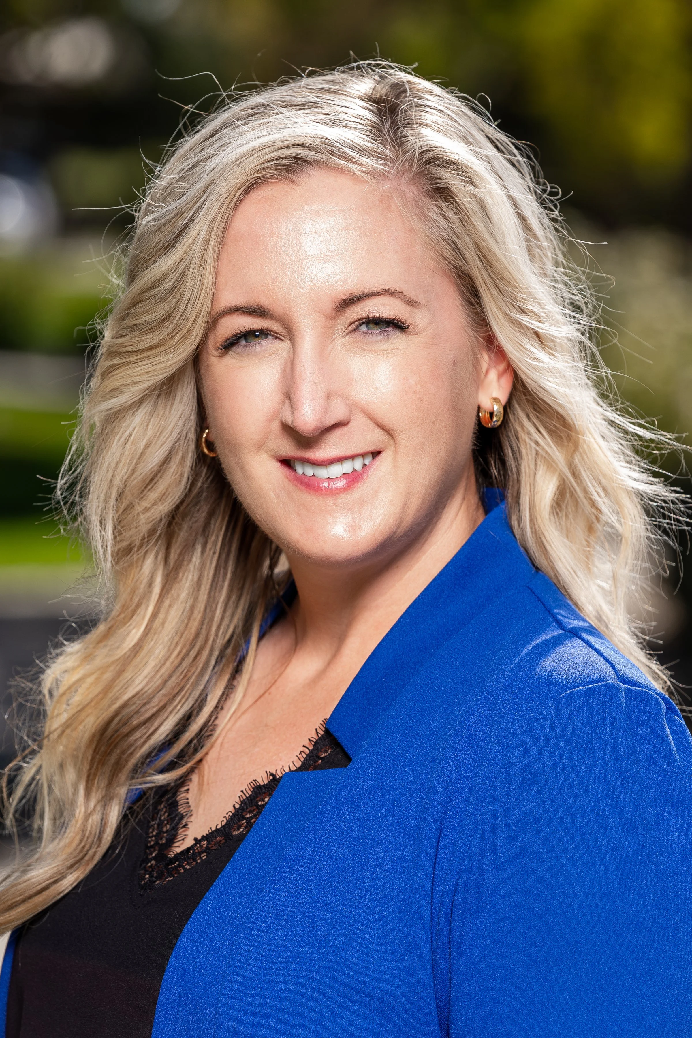 A woman with long blonde hair, wearing a bright blue blazer and black top, smiling outdoors with blurred greenery in the background.