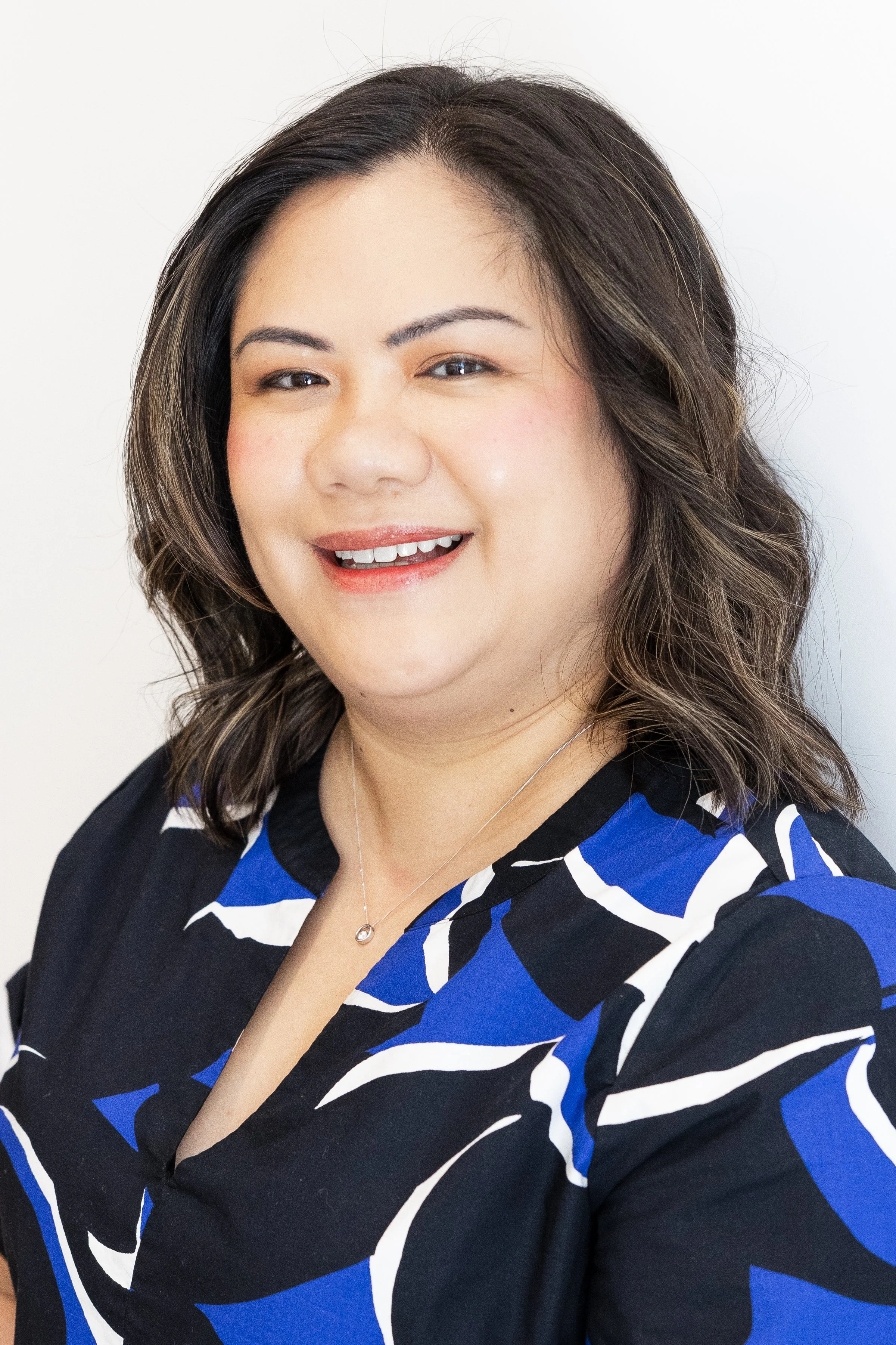 A woman with medium-length dark brown hair styled in loose waves, wearing a black blouse with blue and white abstract patterns, a small pendant necklace, and smiling at the camera against a plain white background.