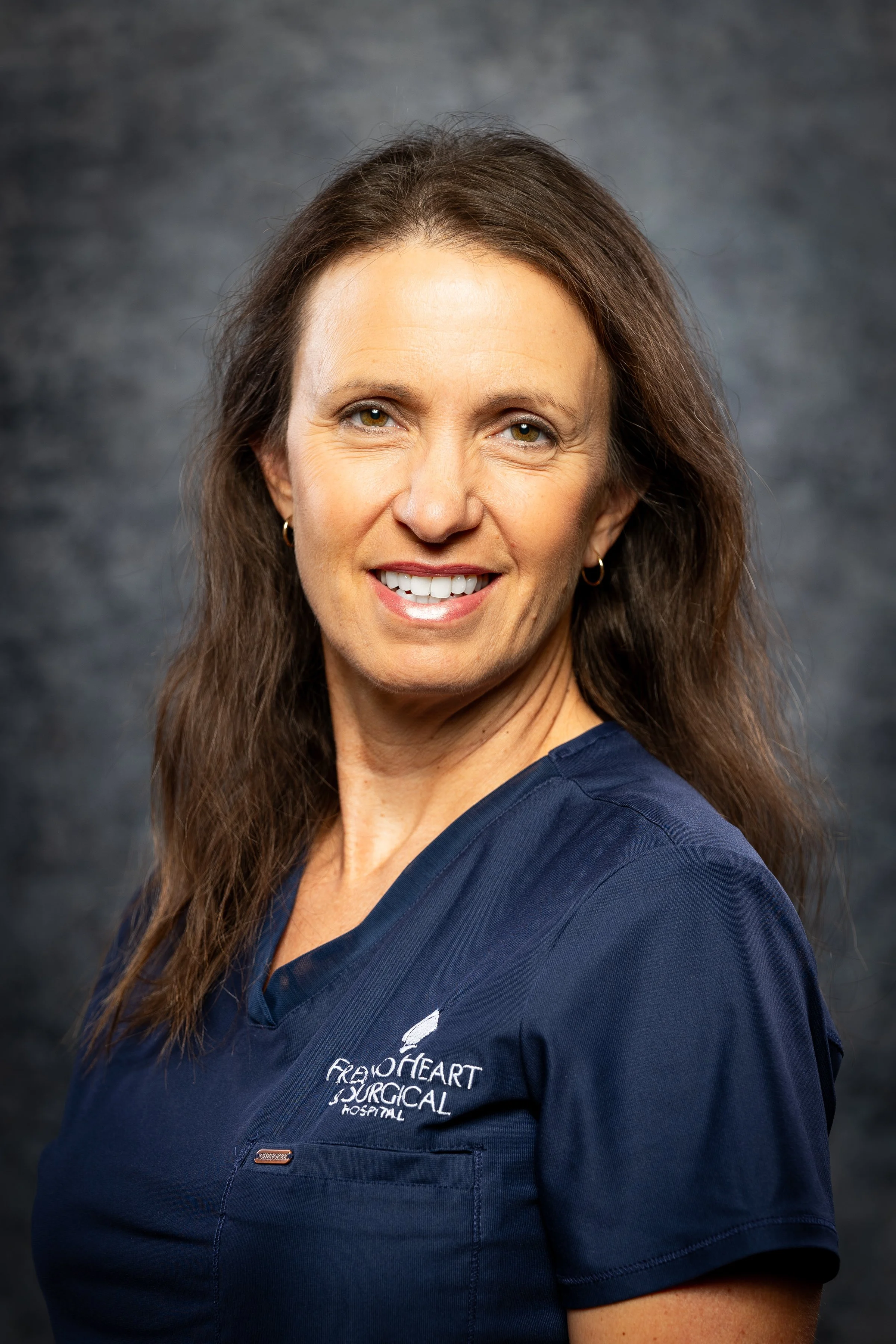 A woman with shoulder-length brown hair smiling, wearing navy scrubs with a logo that reads 'Freo Heart Surgical Hospital,' against a dark blurred background.
