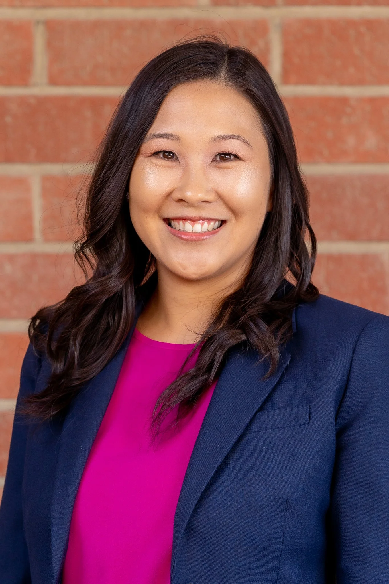 A woman with dark wavy hair smiling, wearing a navy blazer over a magenta top, standing in front of a brick wall.