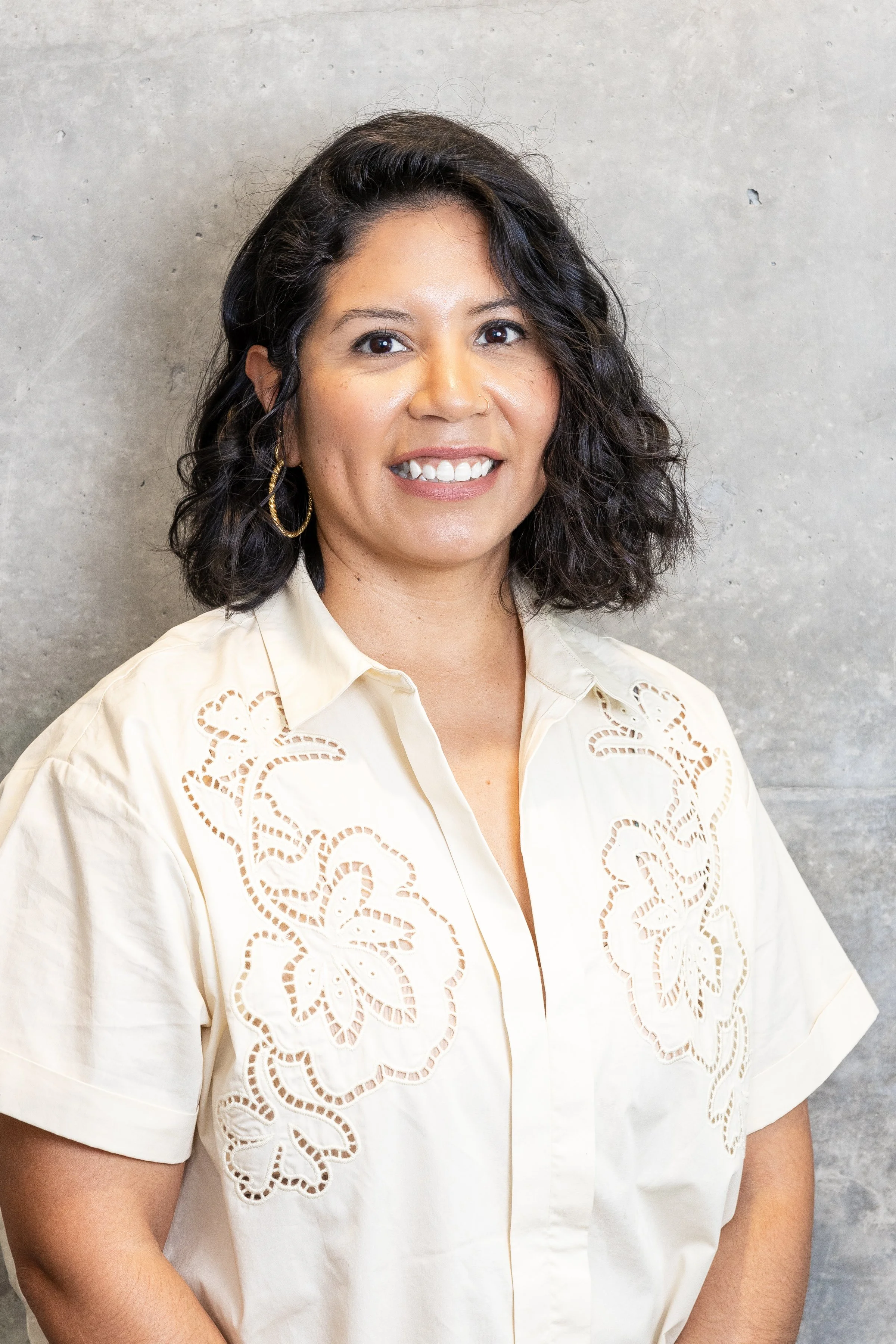 Woman with shoulder-length curly dark hair, wearing a cream-colored blouse with floral cutouts, jewelry, posed against a concrete wall.