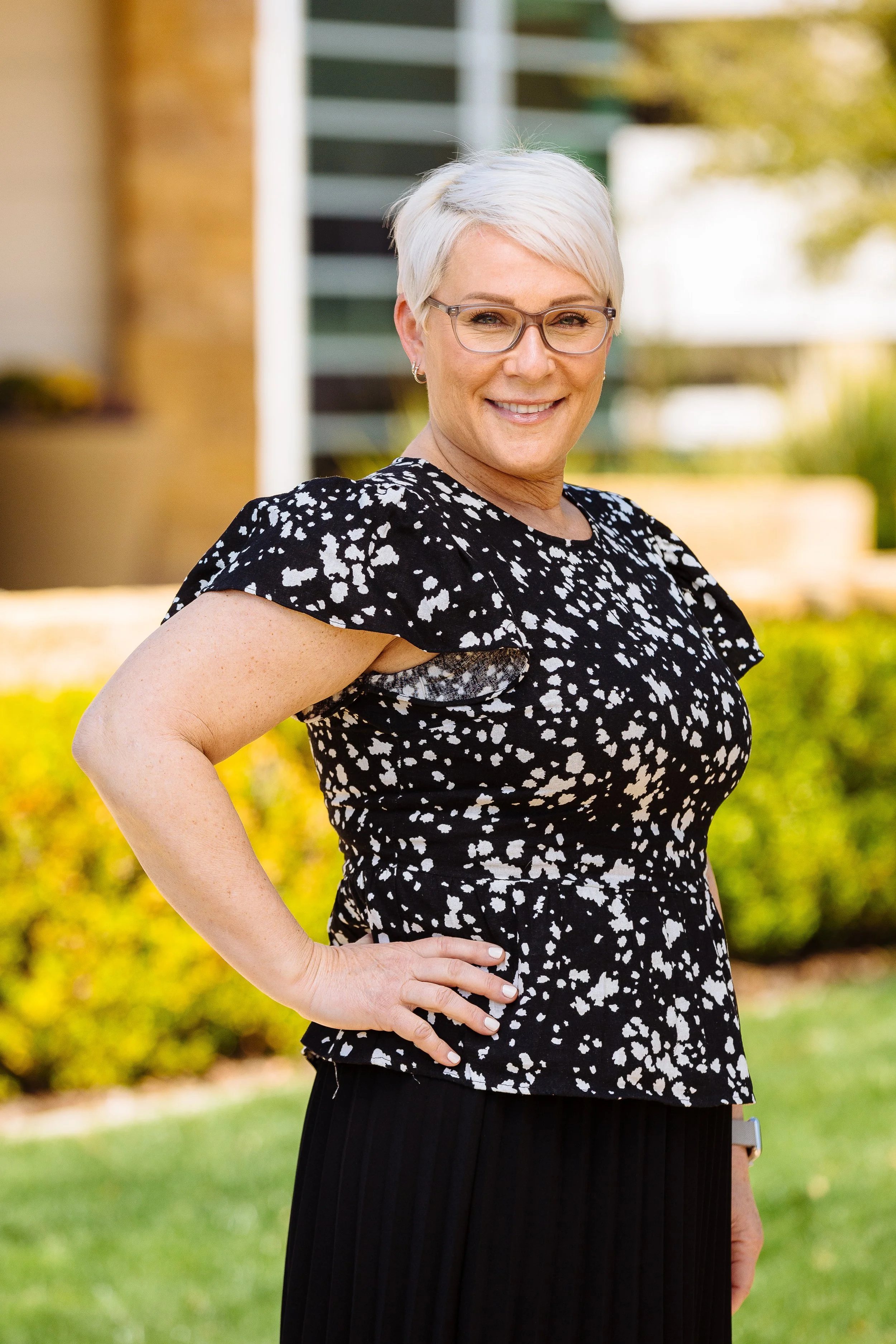 A woman with short white hair and glasses, smiling, standing outside in front of a modern building and greenery.