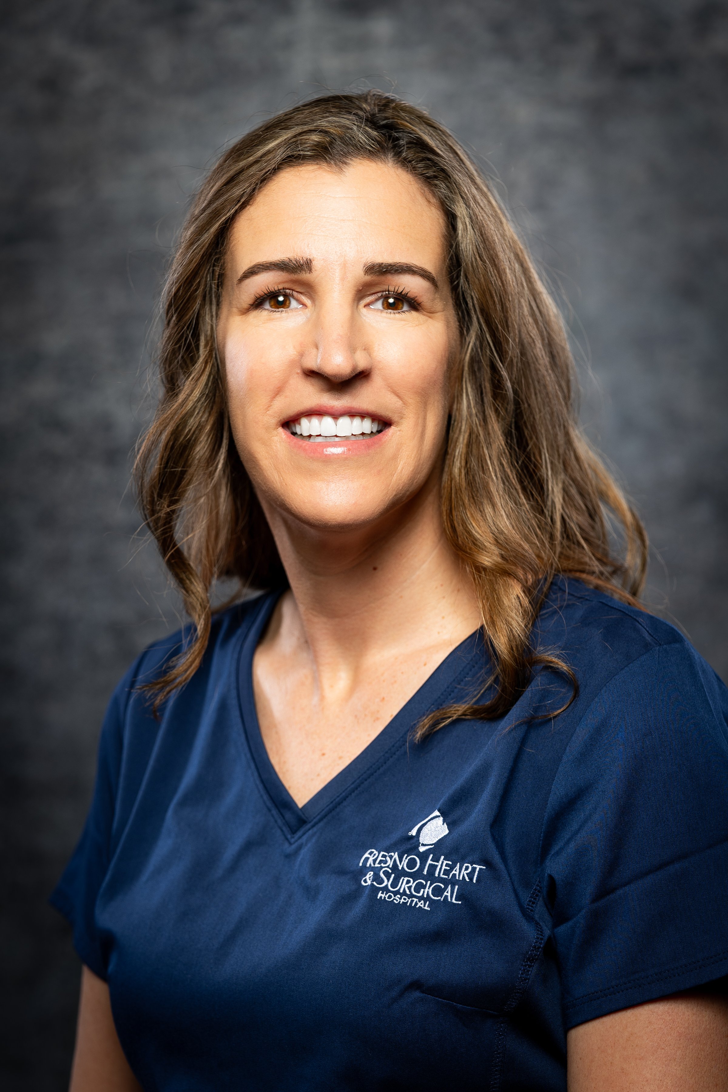 A woman with shoulder-length brown hair smiling, wearing navy blue scrubs with 'Fresno Heart & Surgical Hospital' logo.