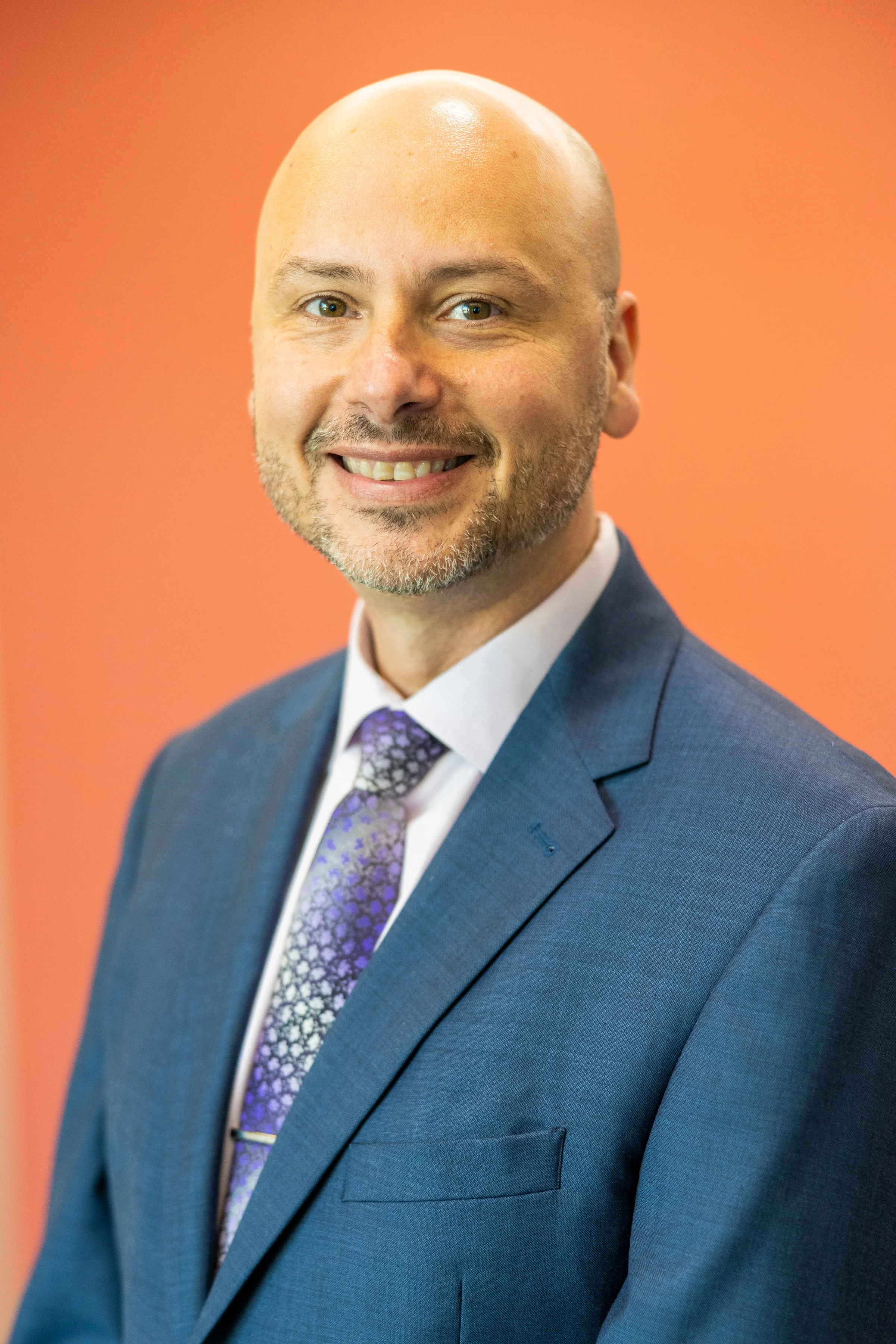 Professional portrait of a smiling man with a bald head, light beard, wearing a blue suit, white shirt, and patterned purple tie, against a coral background.