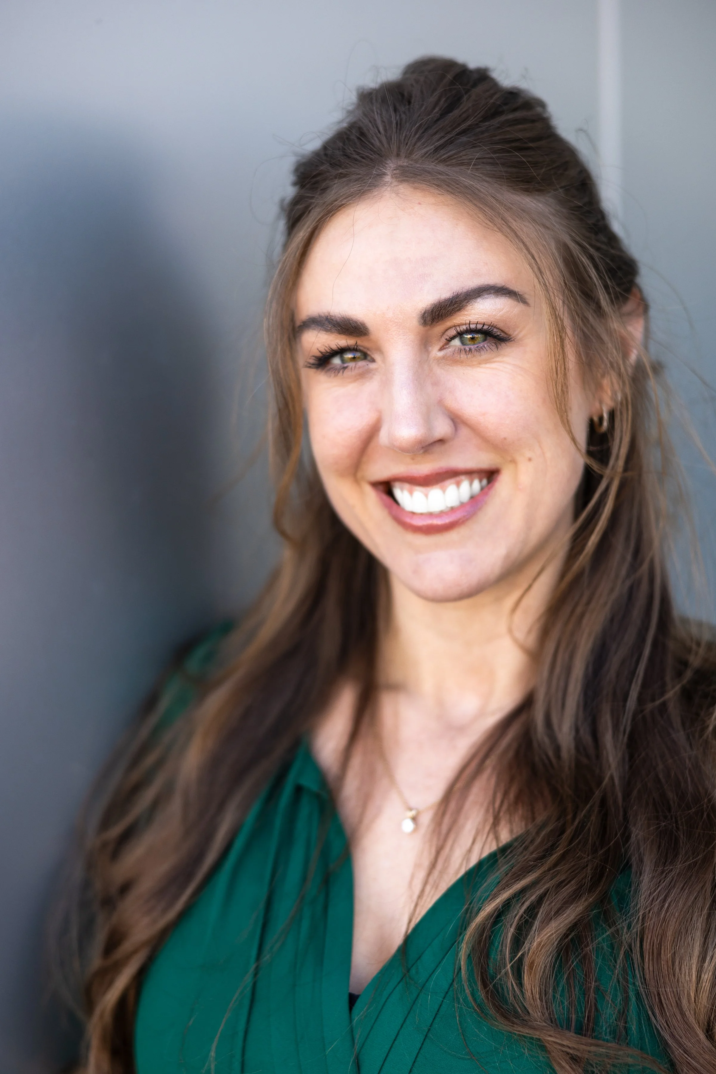 A smiling woman with long brown hair, green eyes, and earrings, wearing a green top and a necklace, leaning against a gray wall.