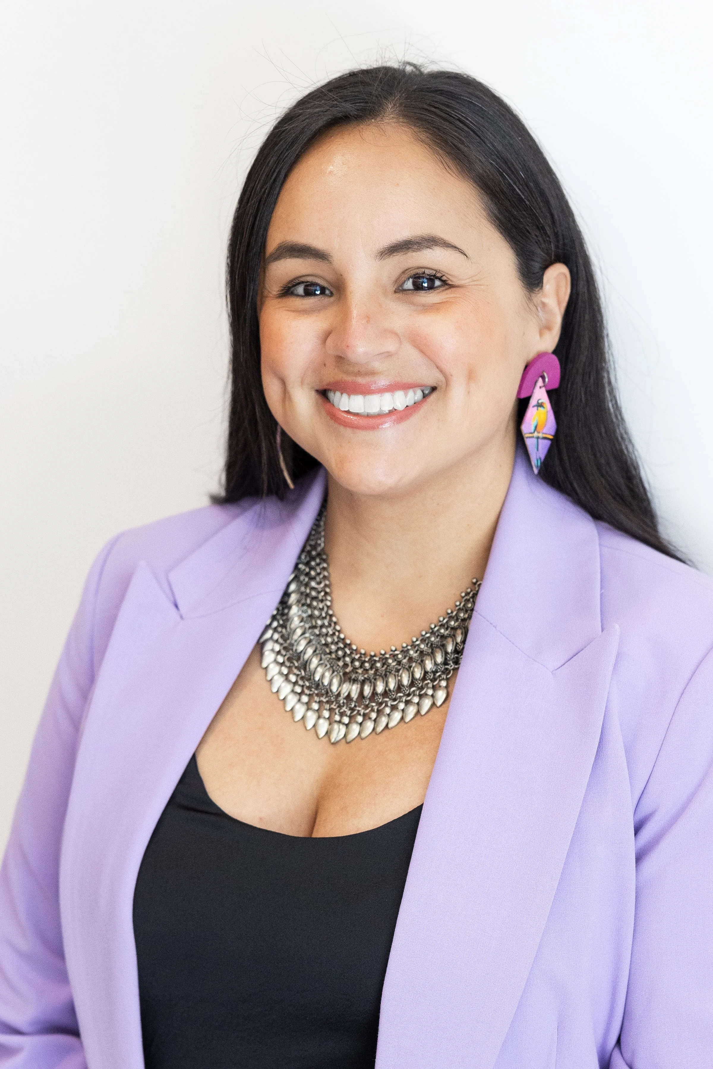 A woman with long dark hair, smiling, wearing a light purple blazer, a black top, and a large silver statement necklace, with colorful earrings featuring a toucan, standing against a plain white background.