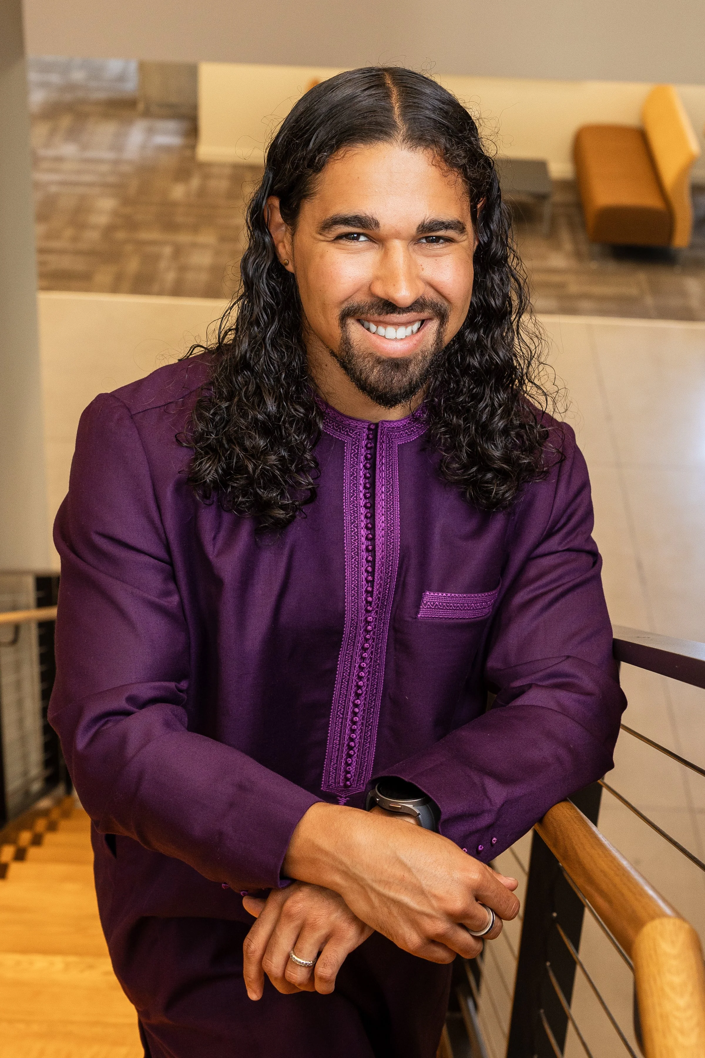 Man with long curly hair and a goatee wearing a purple traditional shirt, smiling, leaning on a railing in a modern indoor setting.