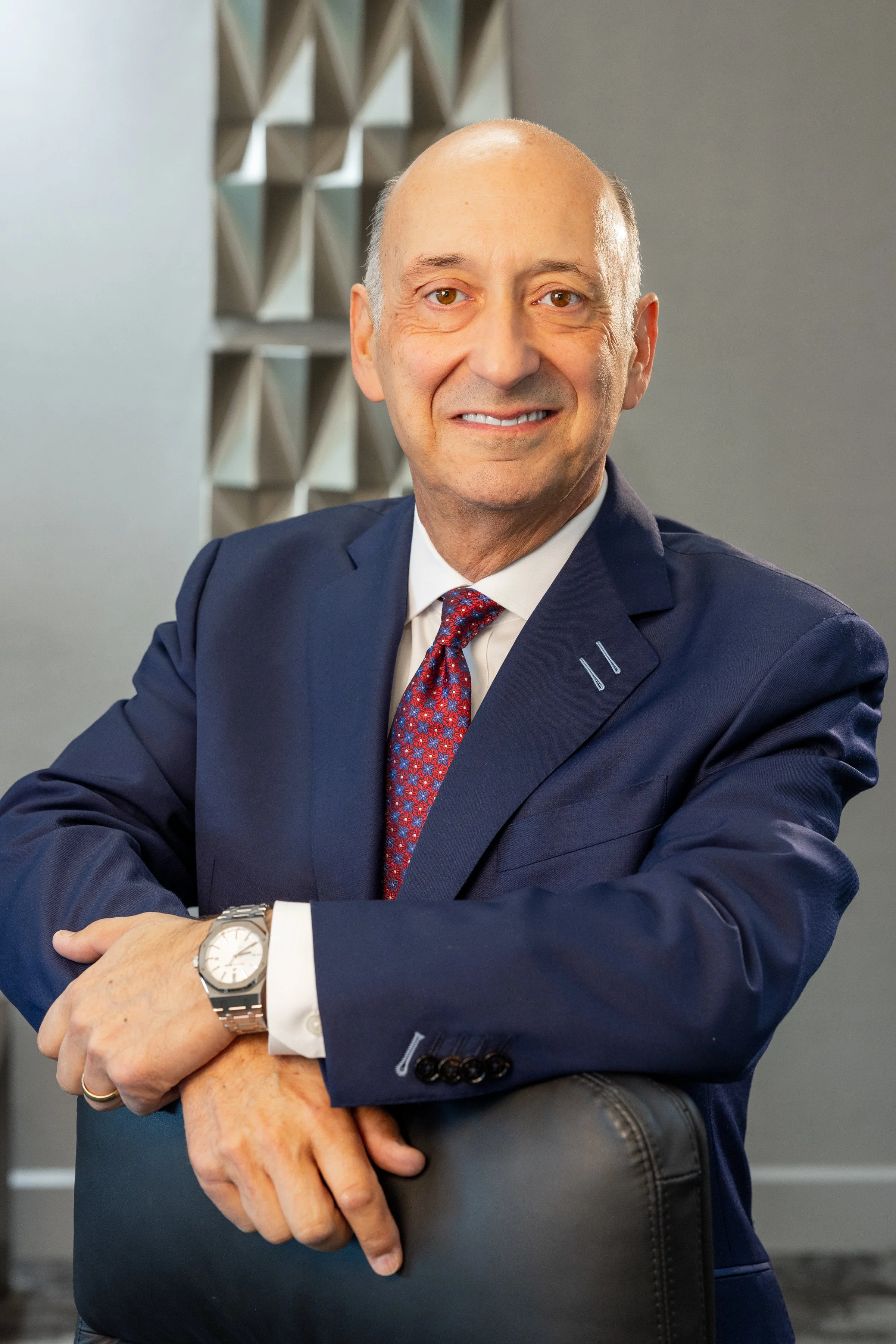A middle-aged man in a navy suit and red patterned tie, smiling, with a watch on his left wrist, sitting in an office environment with a gray wall and geometric wall art in the background.