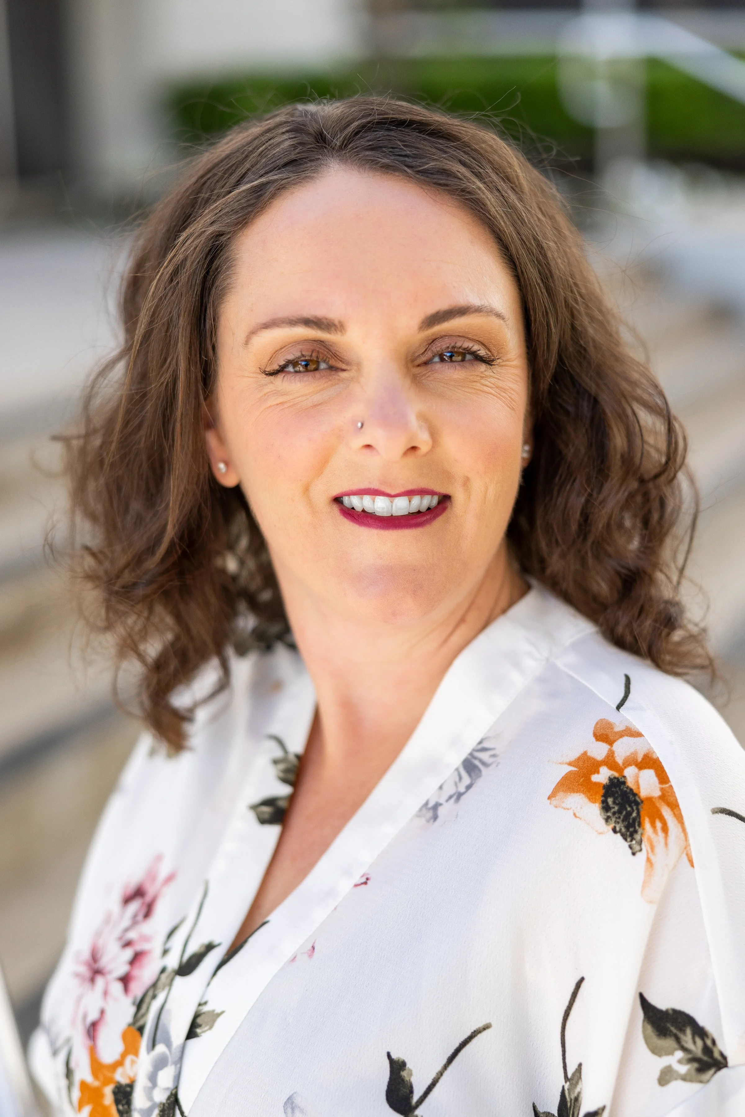 A woman with wavy brown hair wearing a white floral blouse.