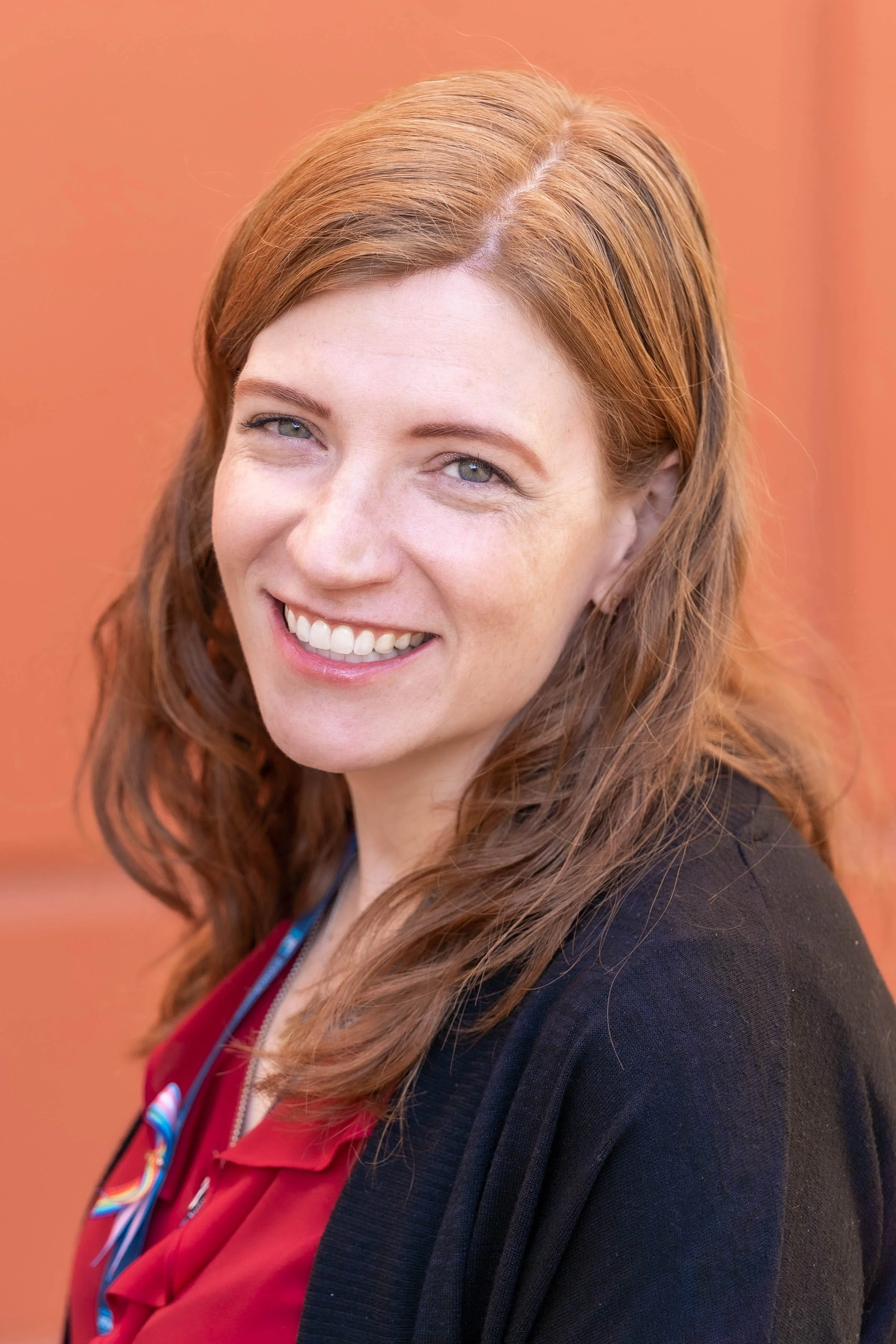 Smiling woman with auburn hair wearing a red top and black cardigan against an orange background.