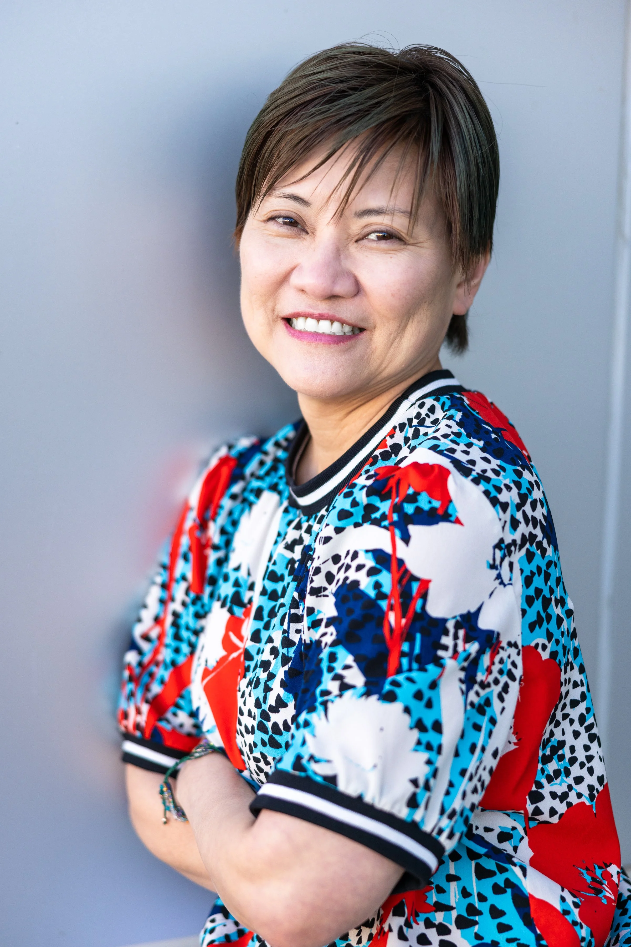 A woman with short brown hair smiling, wearing a colorful patterned shirt with red, white, and blue designs, standing against a plain gray background.