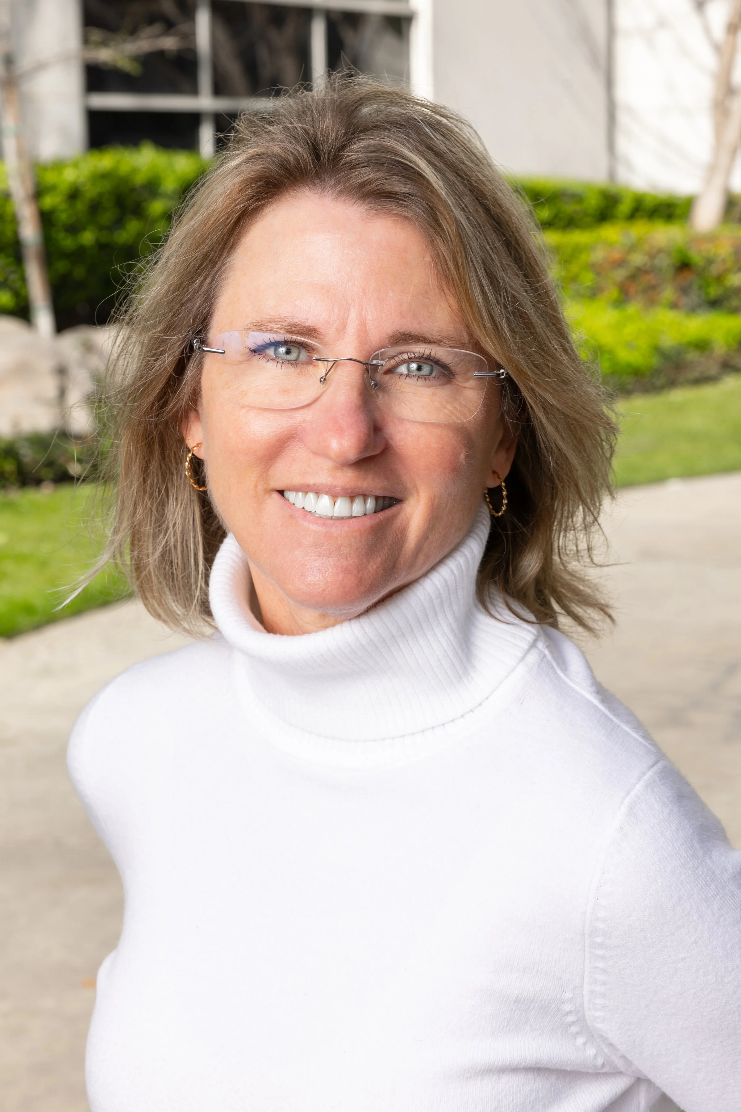 Close-up portrait of a middle-aged woman with shoulder-length light brown hair, wearing glasses and a white turtleneck sweater, smiling outdoors near greenery.