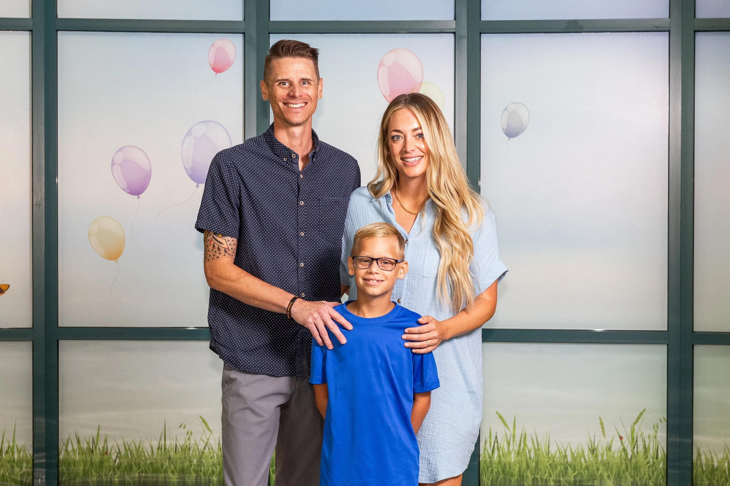 Family of three standing in front of a decorated glass window, with balloons painted on it. The man has short hair, a tattoo sleeve, and is smiling. The woman has long blonde hair and is also smiling. The boy in front has glasses and a blue shirt. Th