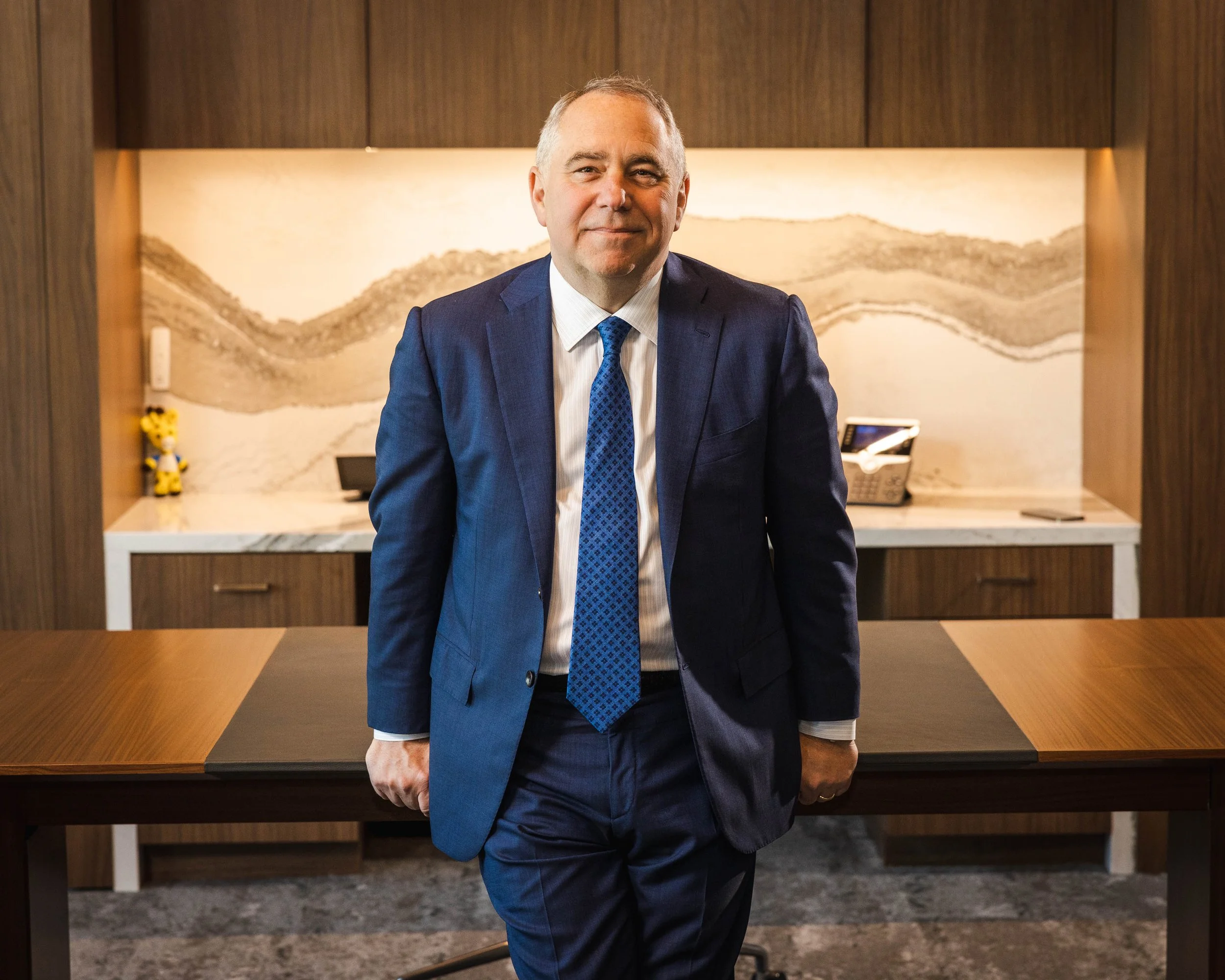 A man in a dark blue suit and blue tie standing behind a wooden desk in a modern office, with a marble wall and abstract artwork in the background.