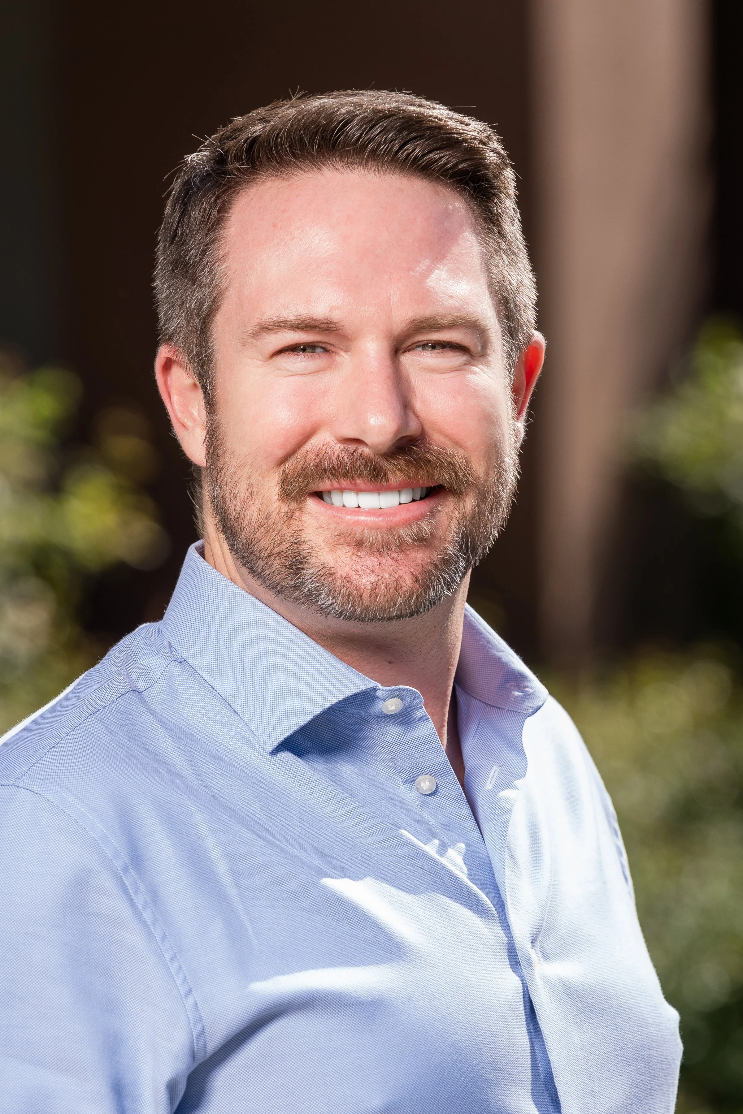A man with short dark hair, a beard, and a mustache, smiling, wearing a light blue dress shirt, outdoors with blurred greenery in the background.
