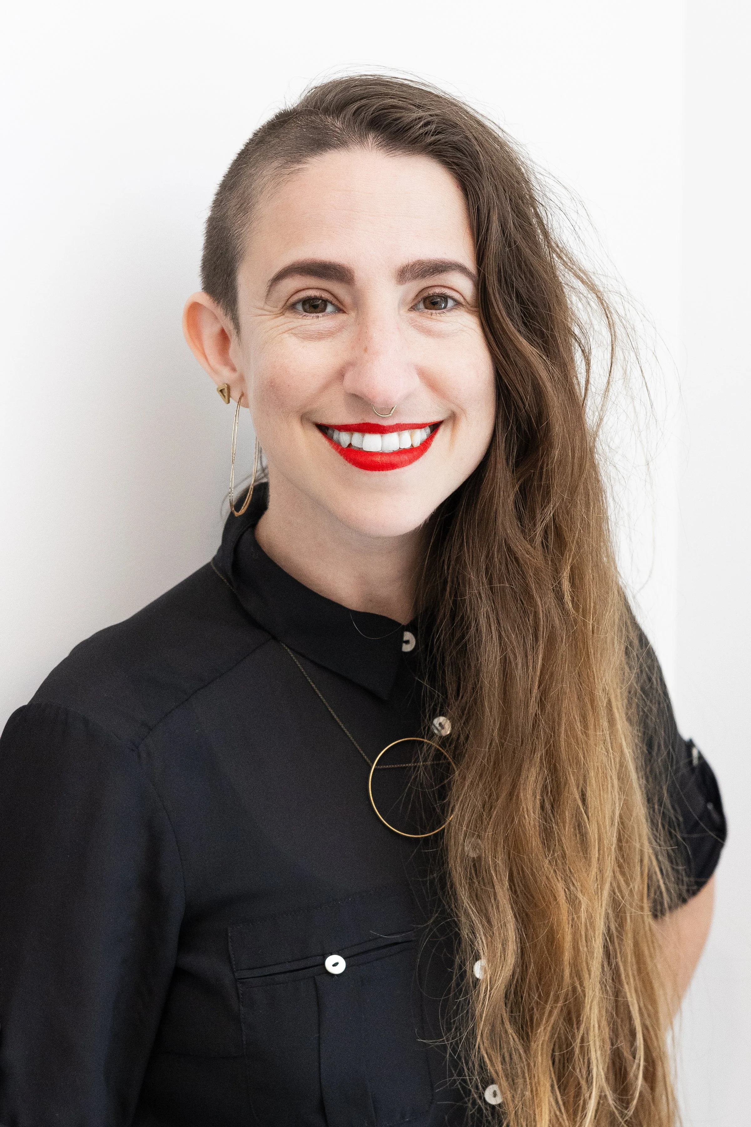 A woman with long brown hair, one side shaved, smiling and wearing red lipstick, gold hoop earrings, and a black shirt with white buttons, standing against a white background.