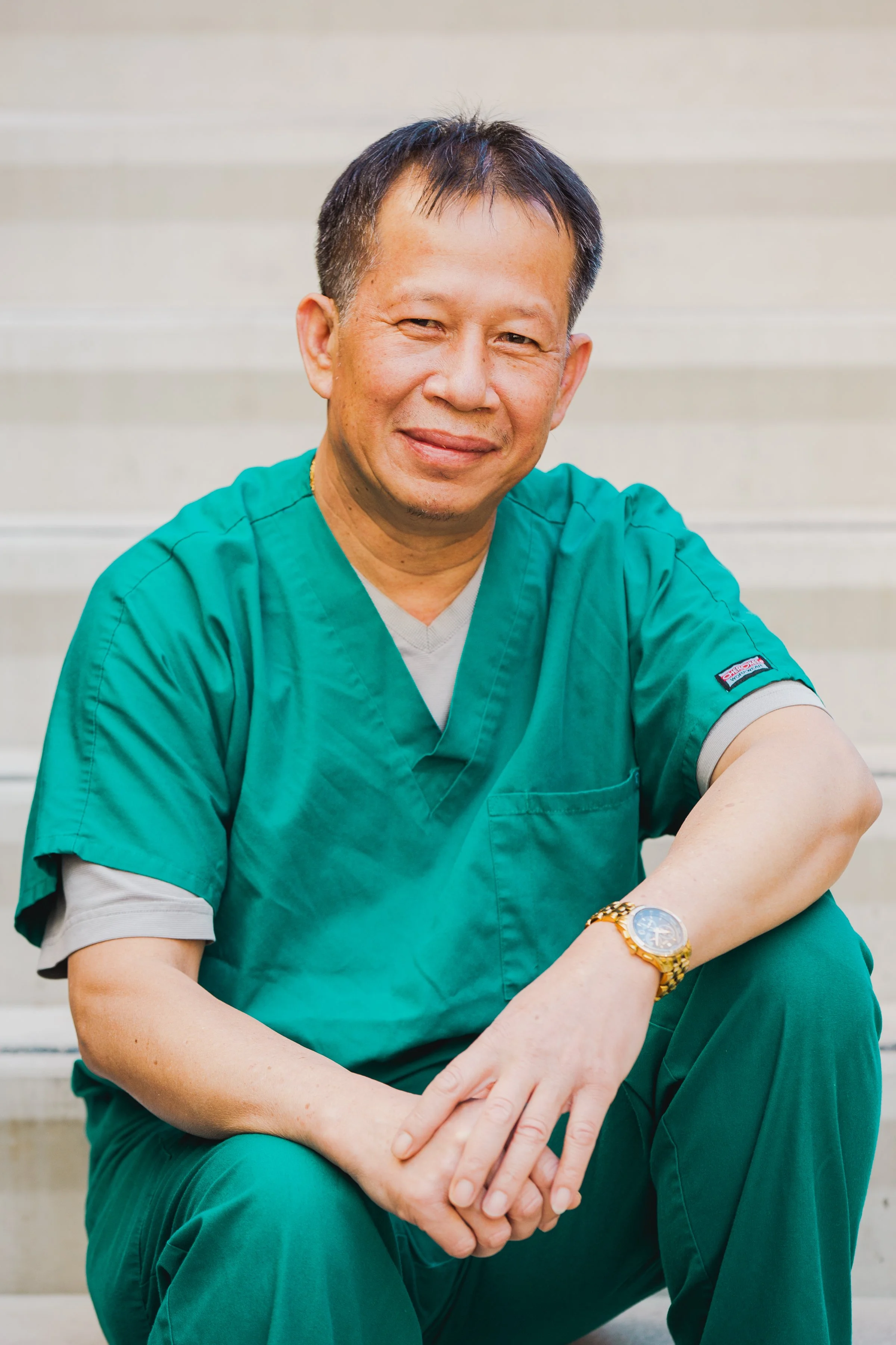 A smiling man in green medical scrubs sitting on stairs with hands clasped, wearing a gold watch.